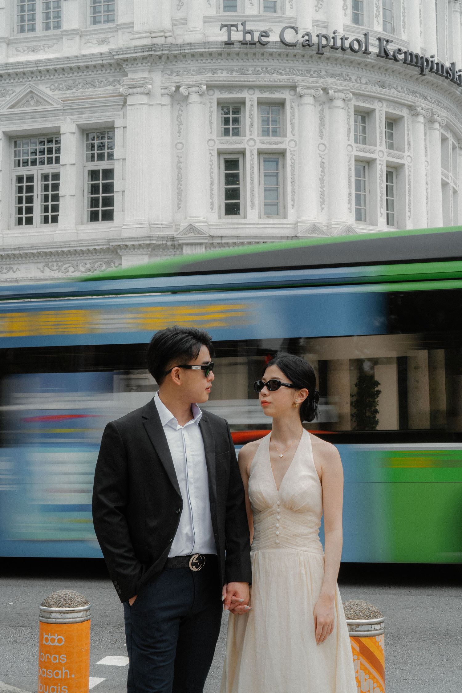 A couple in formal attire poses in front of historic architecture as colorful buses blur past in the background.
