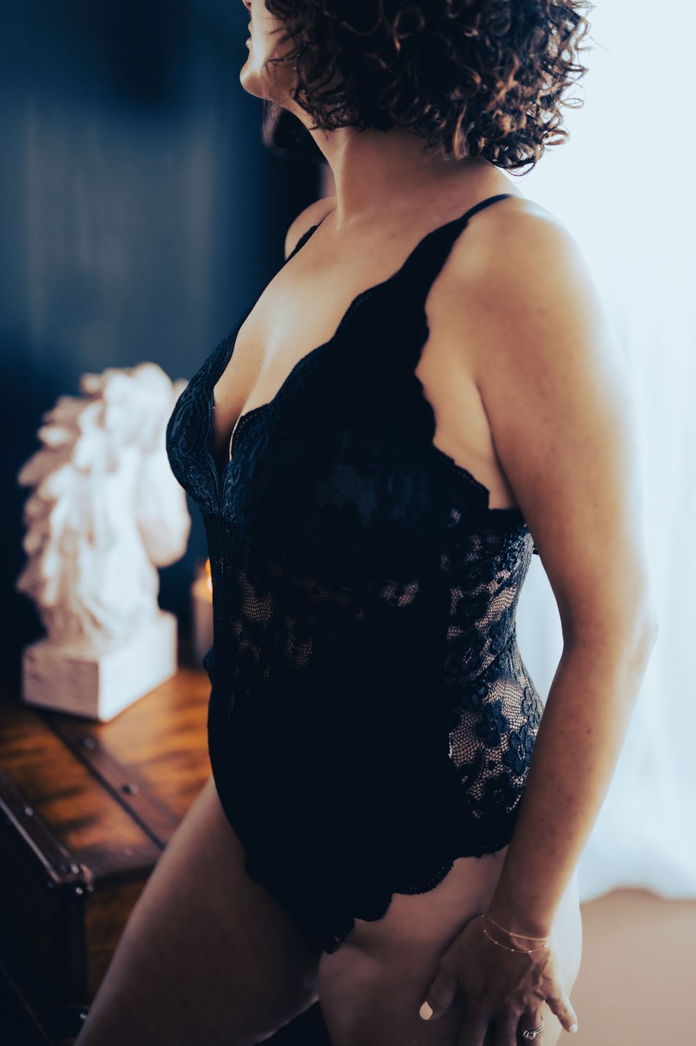 Woman in black lace lingerie bodysuit posing elegantly indoors with soft natural light and decorative statue in background.