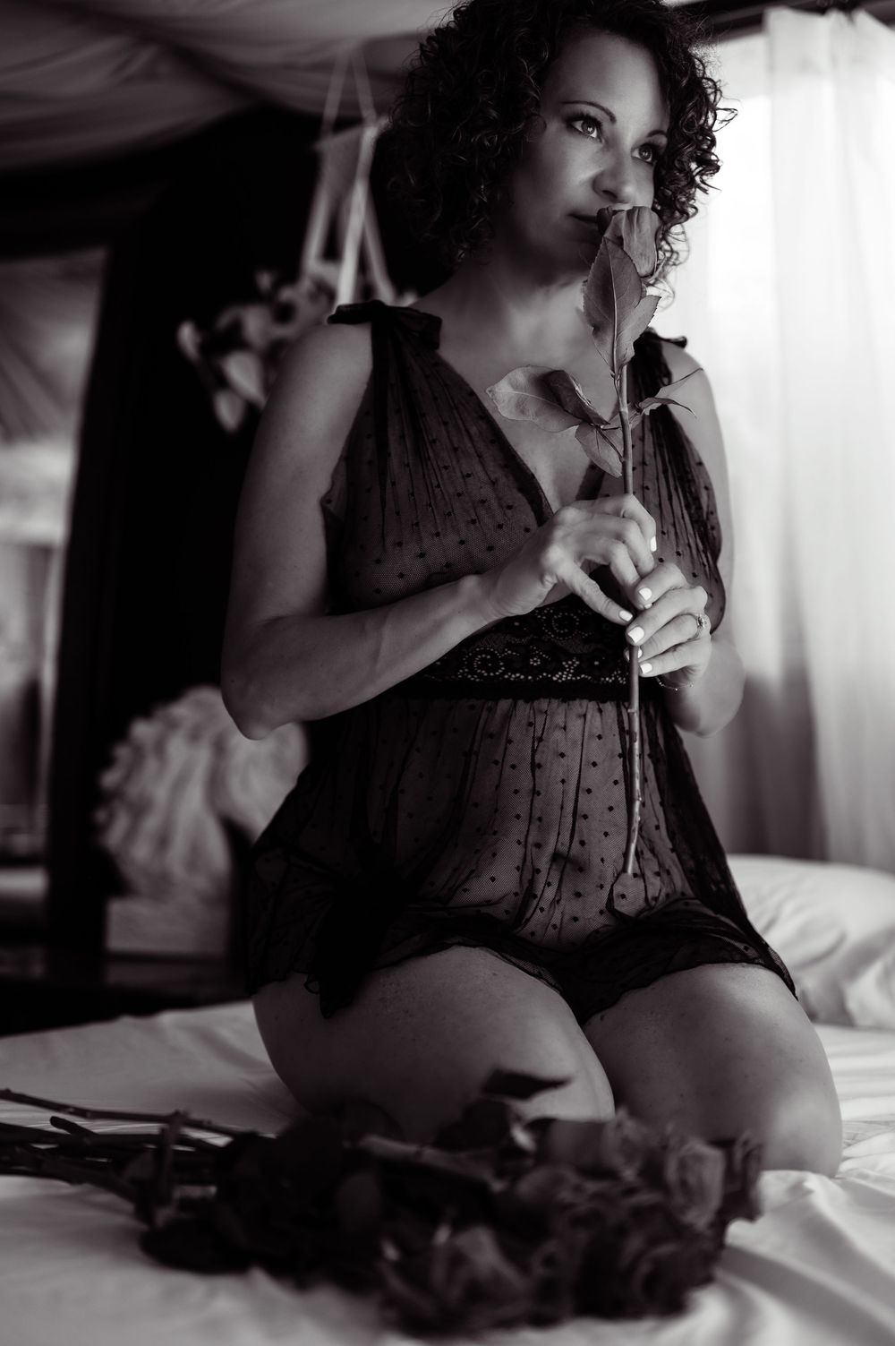 Black and white boudoir photo of a woman in lingerie kneeling on a bed, holding a rose near her face.