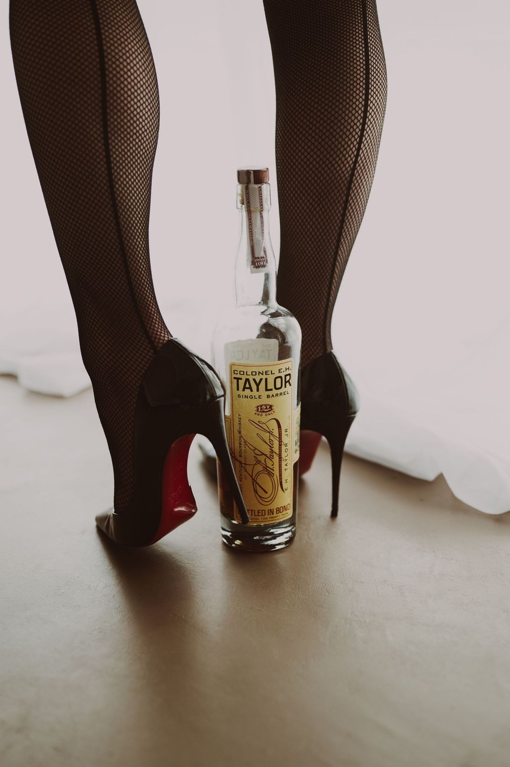A sequence of black fishnet stockings and red high heels next to a bottle of Taylor whiskey against a white wall.
