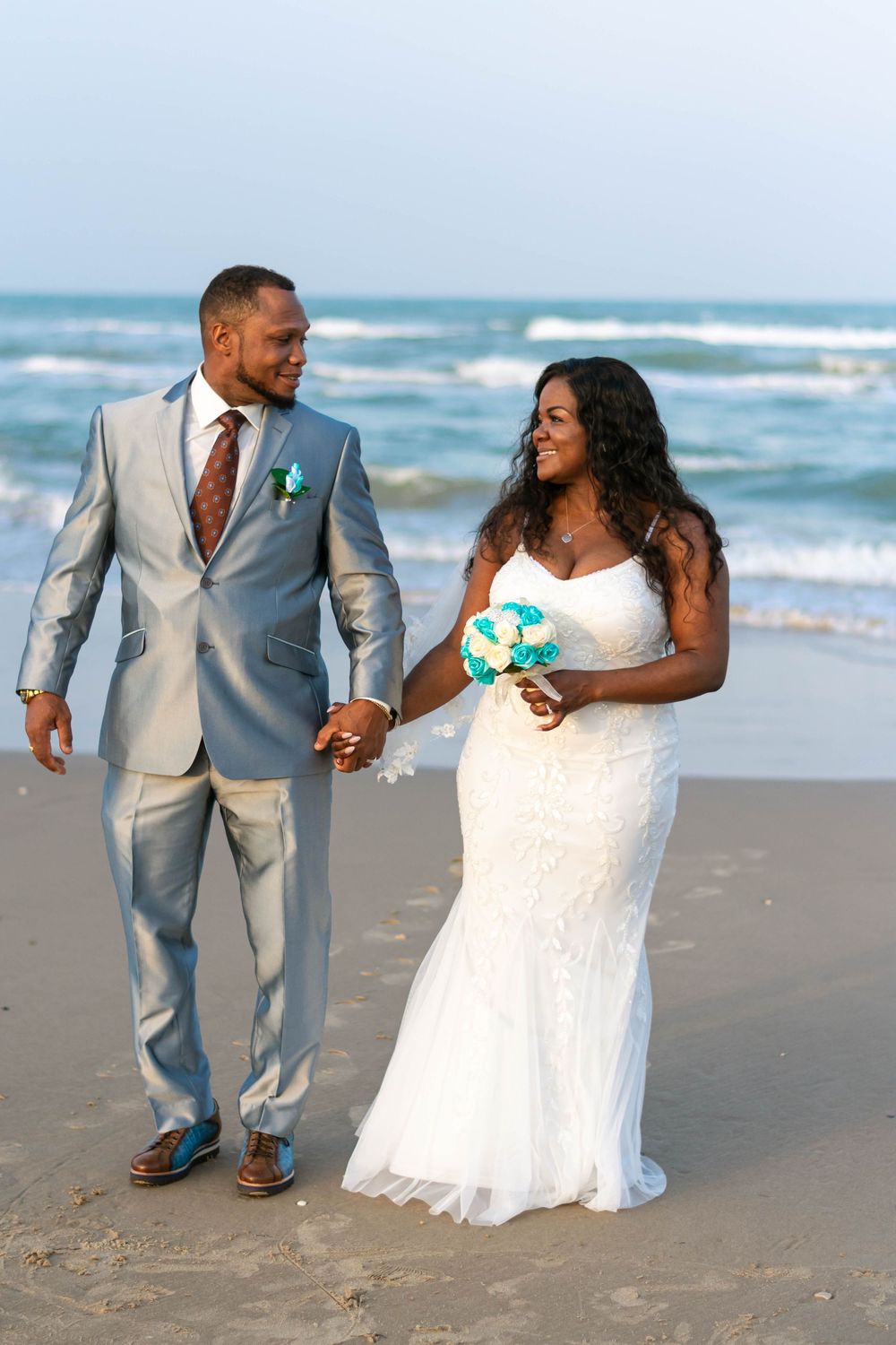 Newlywed couple holding hands on a beach, bride in white lace gown with teal bouquet, groom in gray suit.