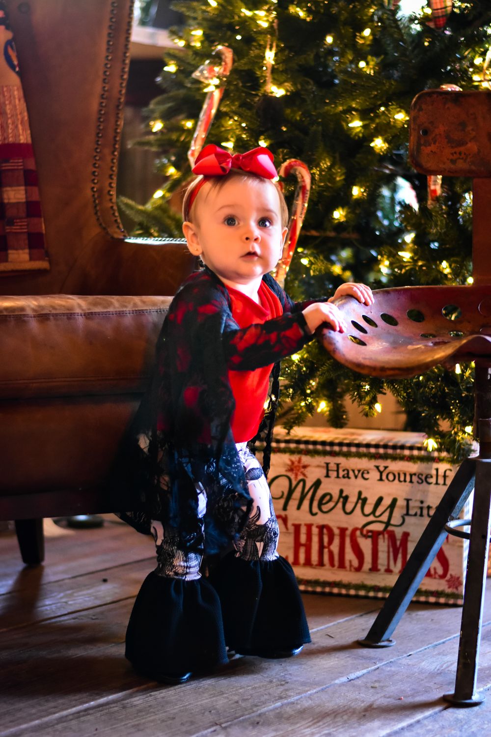 Baby in holiday outfit sitting near Christmas tree and decorative signs.
