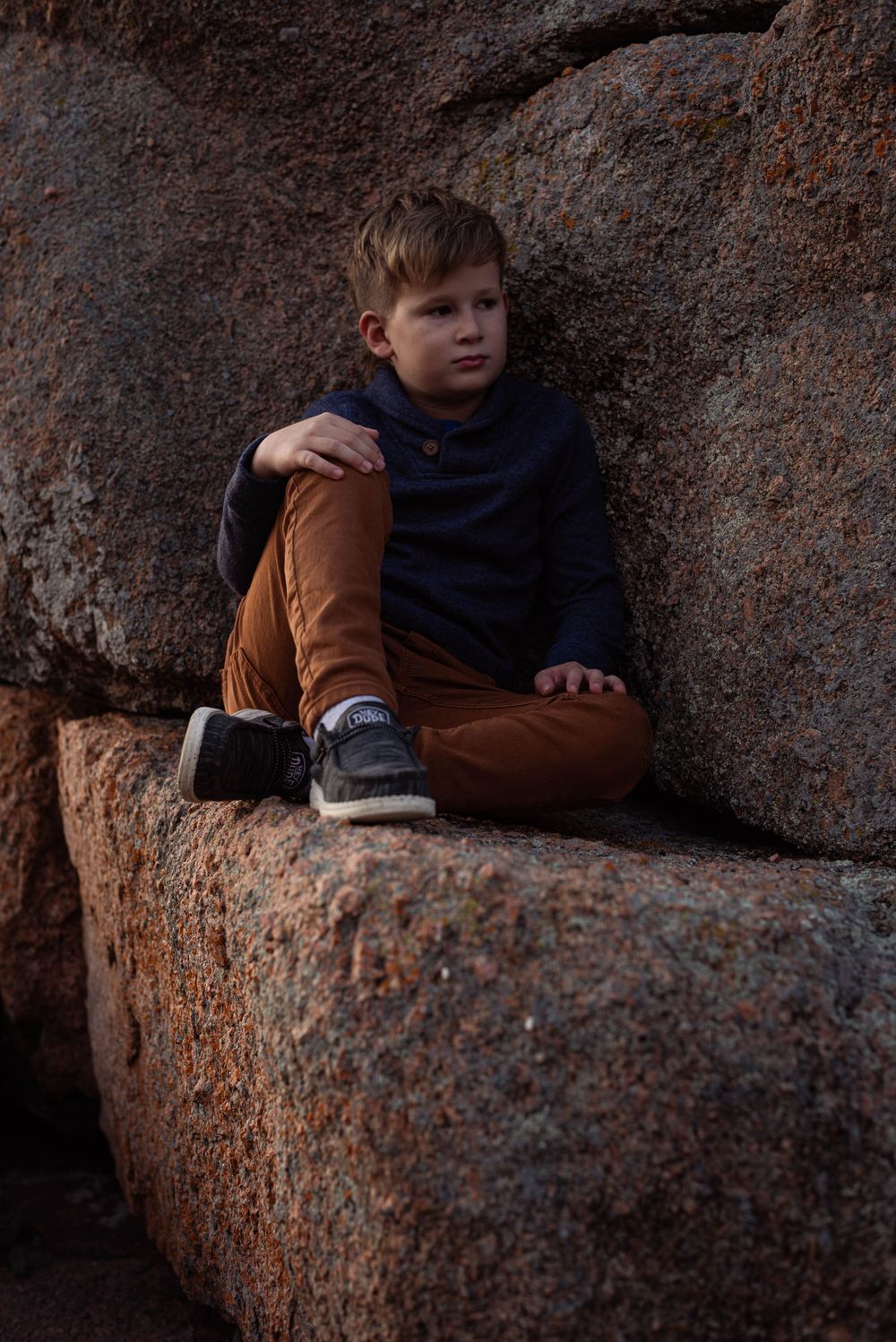 A figure sits nestled in a rocky crevice wearing brown and navy clothing.