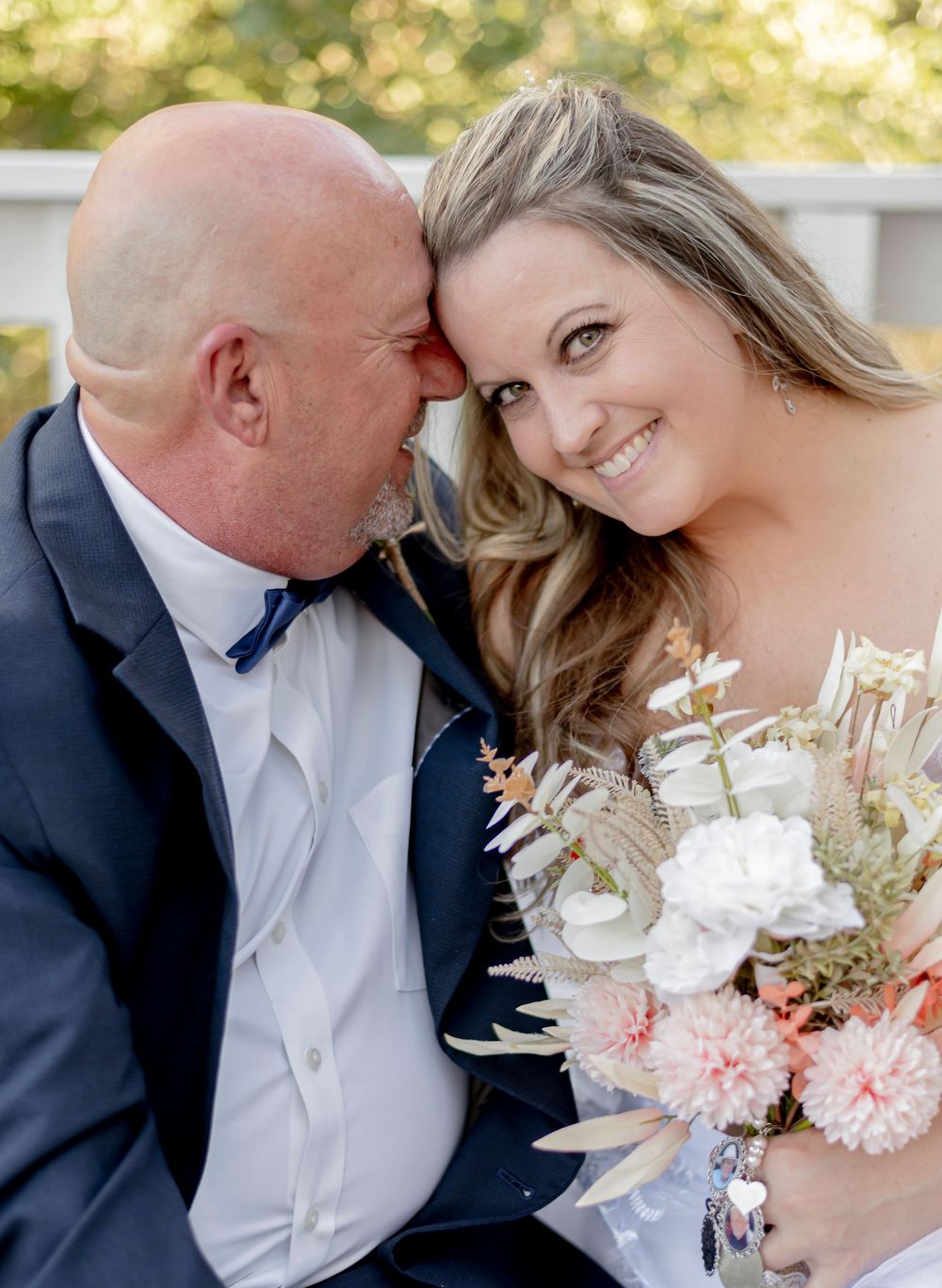 A bride holding a floral bouquet smiles as a bald groom in a navy suit leans in close during their wedding day.