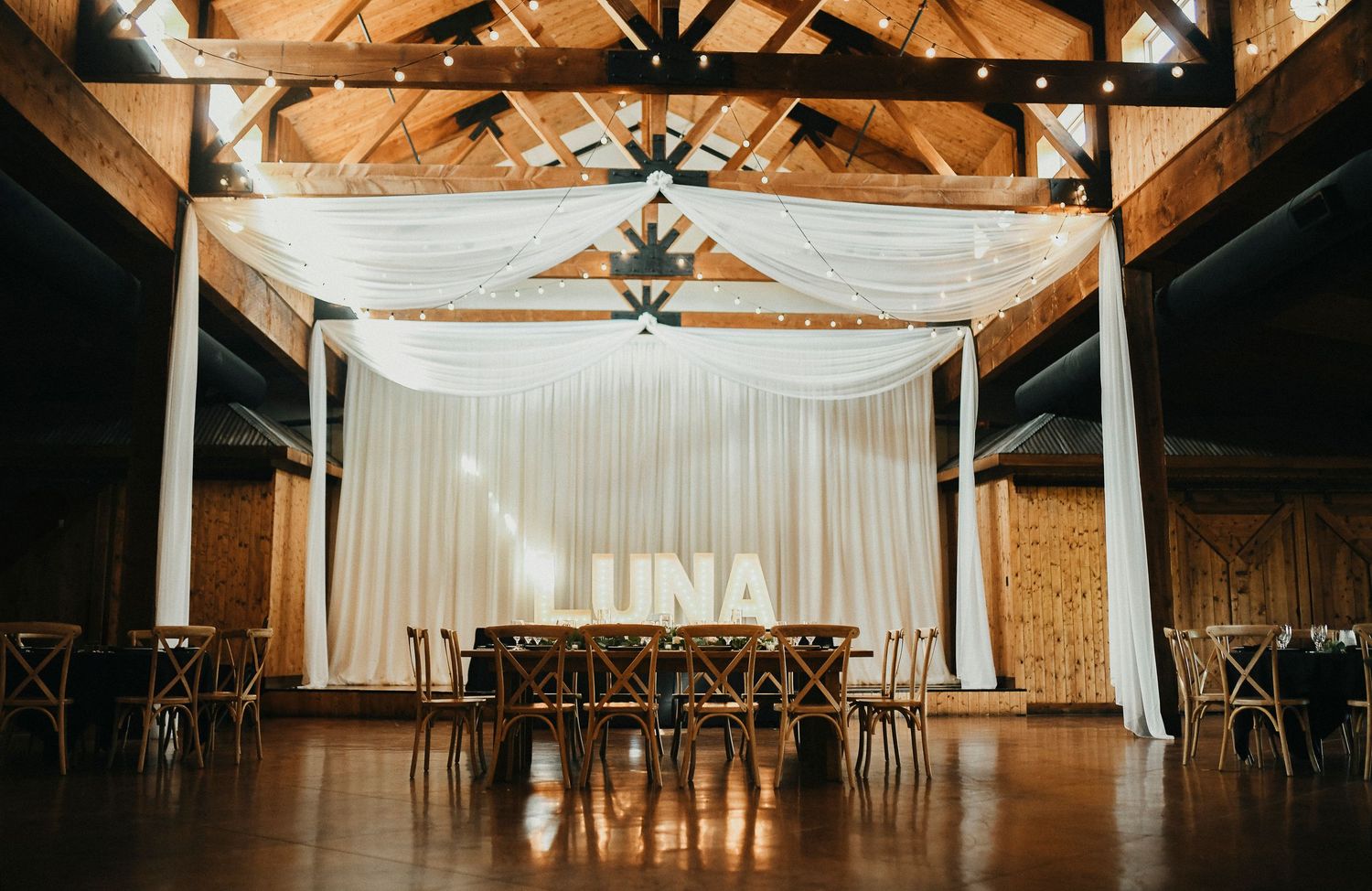 Reception hall at Lucky Spur Ranch featuring flowing white drapery and warm Edison bulb string lights hanging from exposed wooden beams.