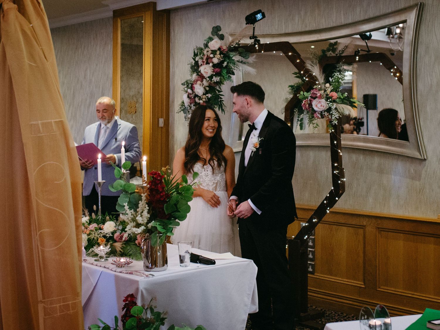 A wedding ceremony takes place under a decorated floral arch in an intimate indoor venue with string lights.