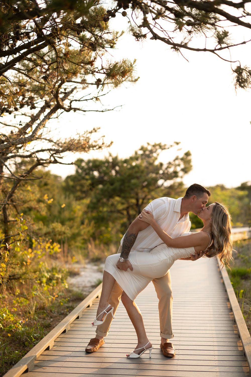 A couple shares a romantic moment while dancing on a wooden boardwalk during golden hour with trees in the background.