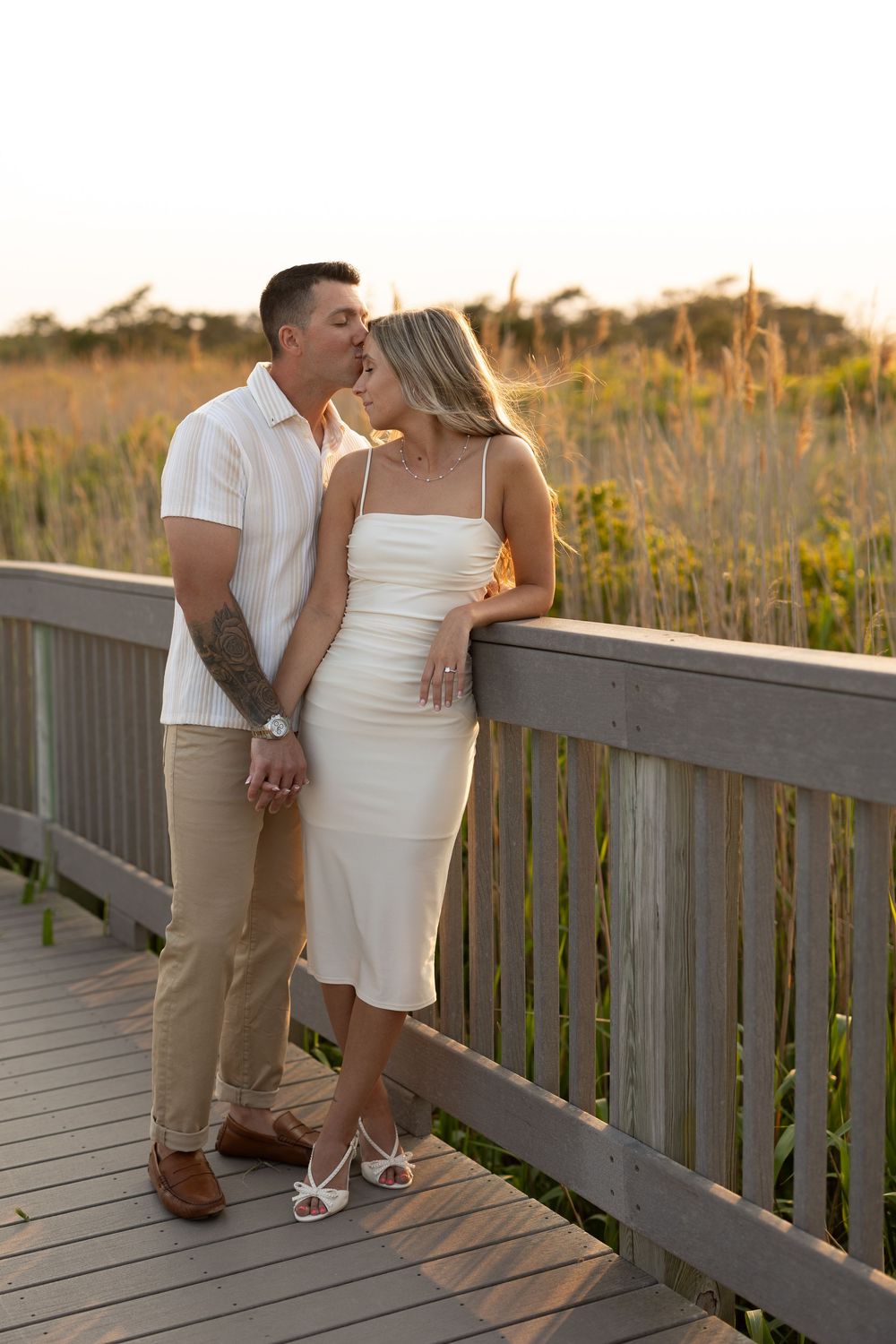 A couple in white and khaki attire share an intimate moment on a wooden boardwalk during golden hour.