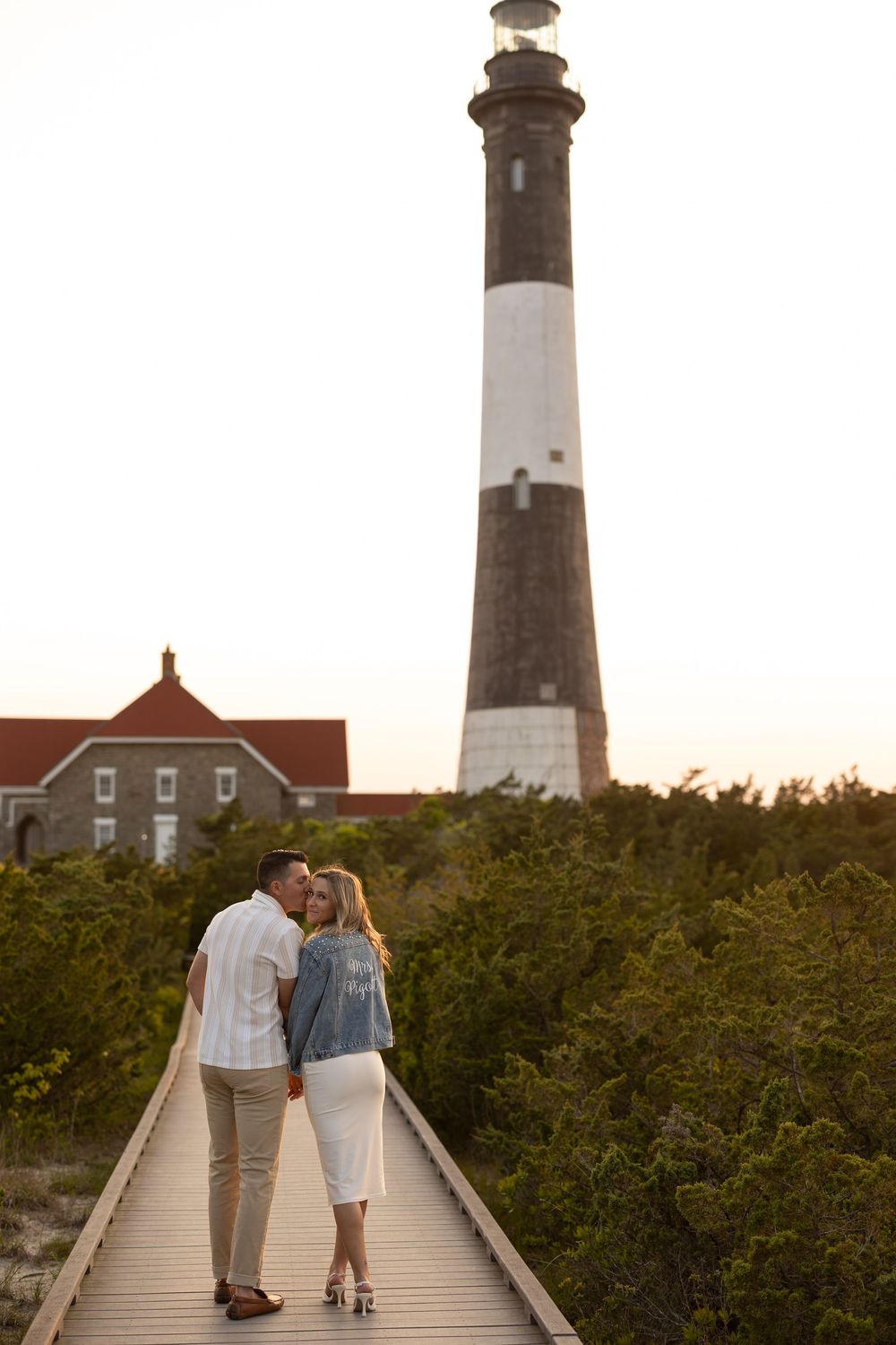 Couple walking on wooden boardwalk path at sunset with Fire Island Lighthouse in the background.