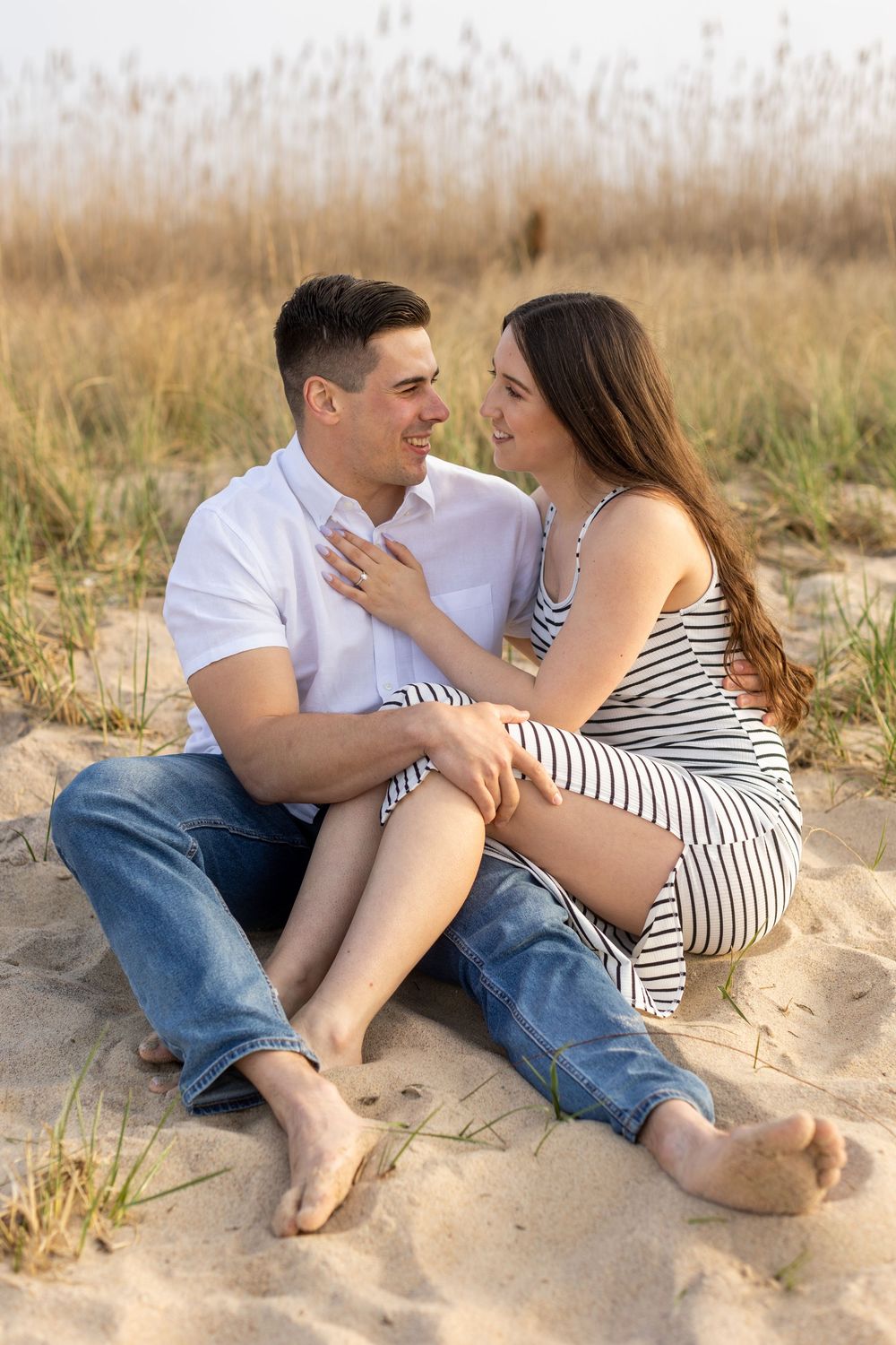 Couple relaxing together on sandy beach dunes with casual attire and playful poses.