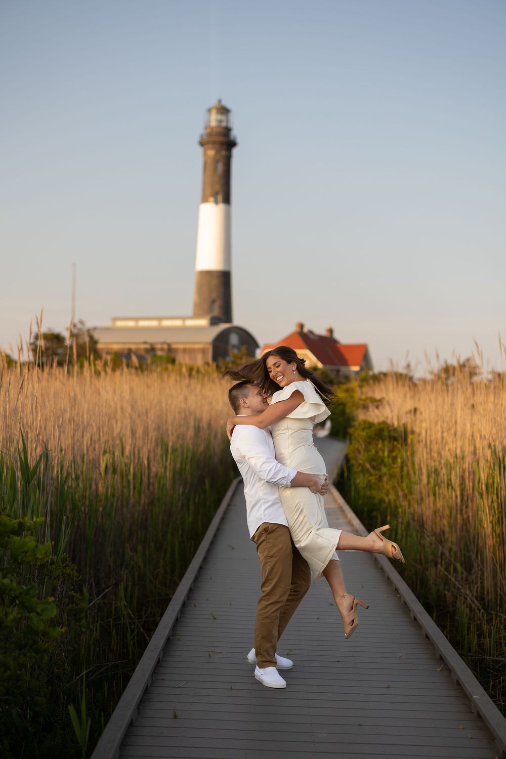 A couple embraces playfully on a wooden boardwalk with a tall lighthouse and coastal vegetation in the background.