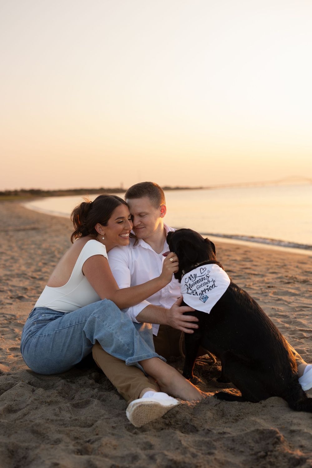 A couple sits together with their black dog on a sandy beach at sunset sharing a tender moment.