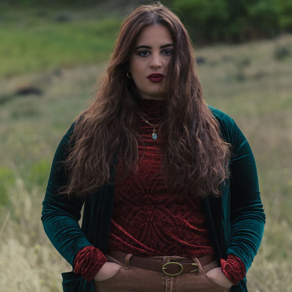 A person with long brown hair poses outdoors wearing a burgundy top and dark teal velvet jacket with a leather belt.