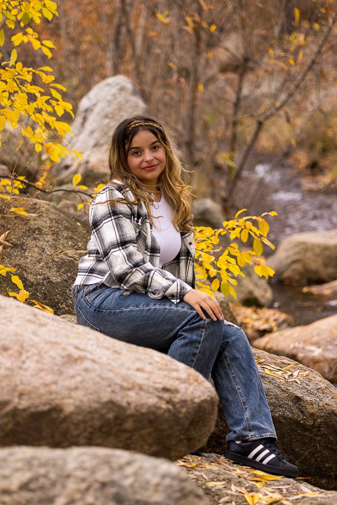 Person in plaid flannel and jeans relaxes on large boulders surrounded by autumn leaves in nature setting.