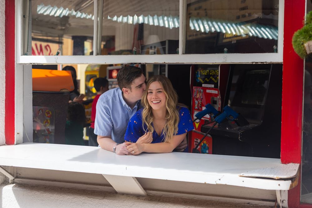 A couple leans together at an outdoor service window with a festive red frame and striped awning above.