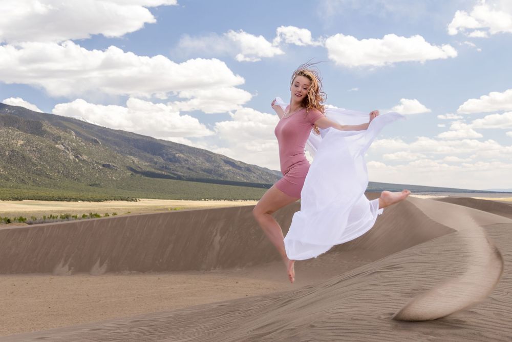 A dancer leaps gracefully on sand dunes against a mountain backdrop under blue skies with white clouds.
