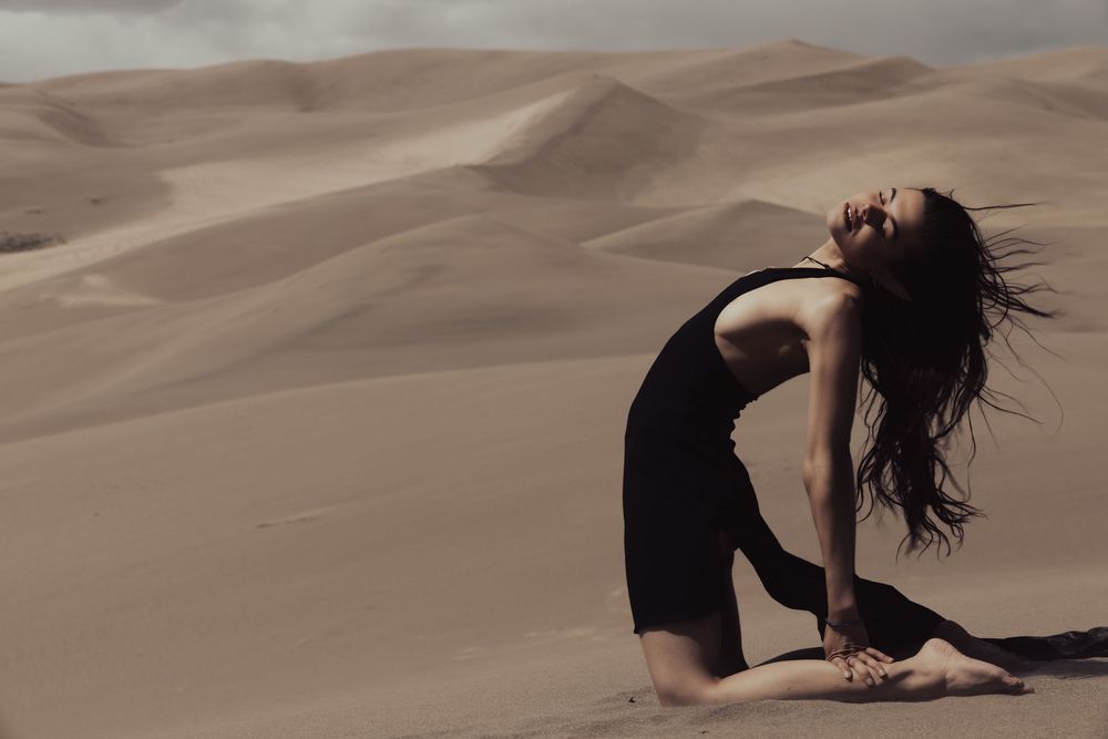 A dancer moves gracefully against a backdrop of sandy desert dunes in a flowing dark dress.
