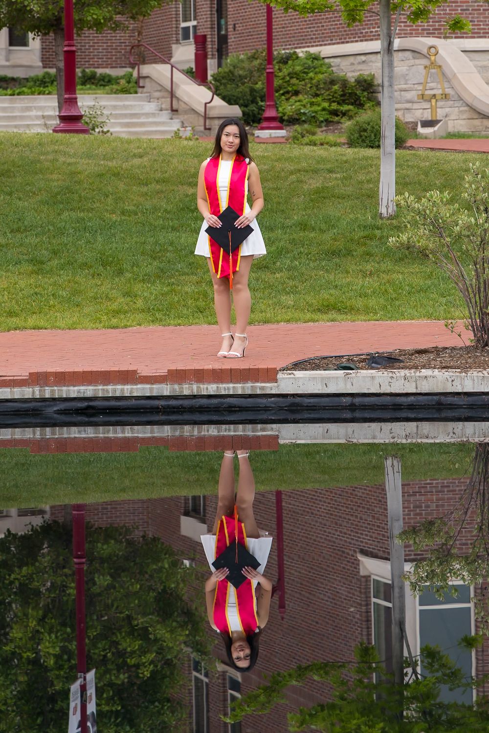 Graduate in a red stole and white dress poses near a reflective pool on a campus with brick buildings and lamp posts.