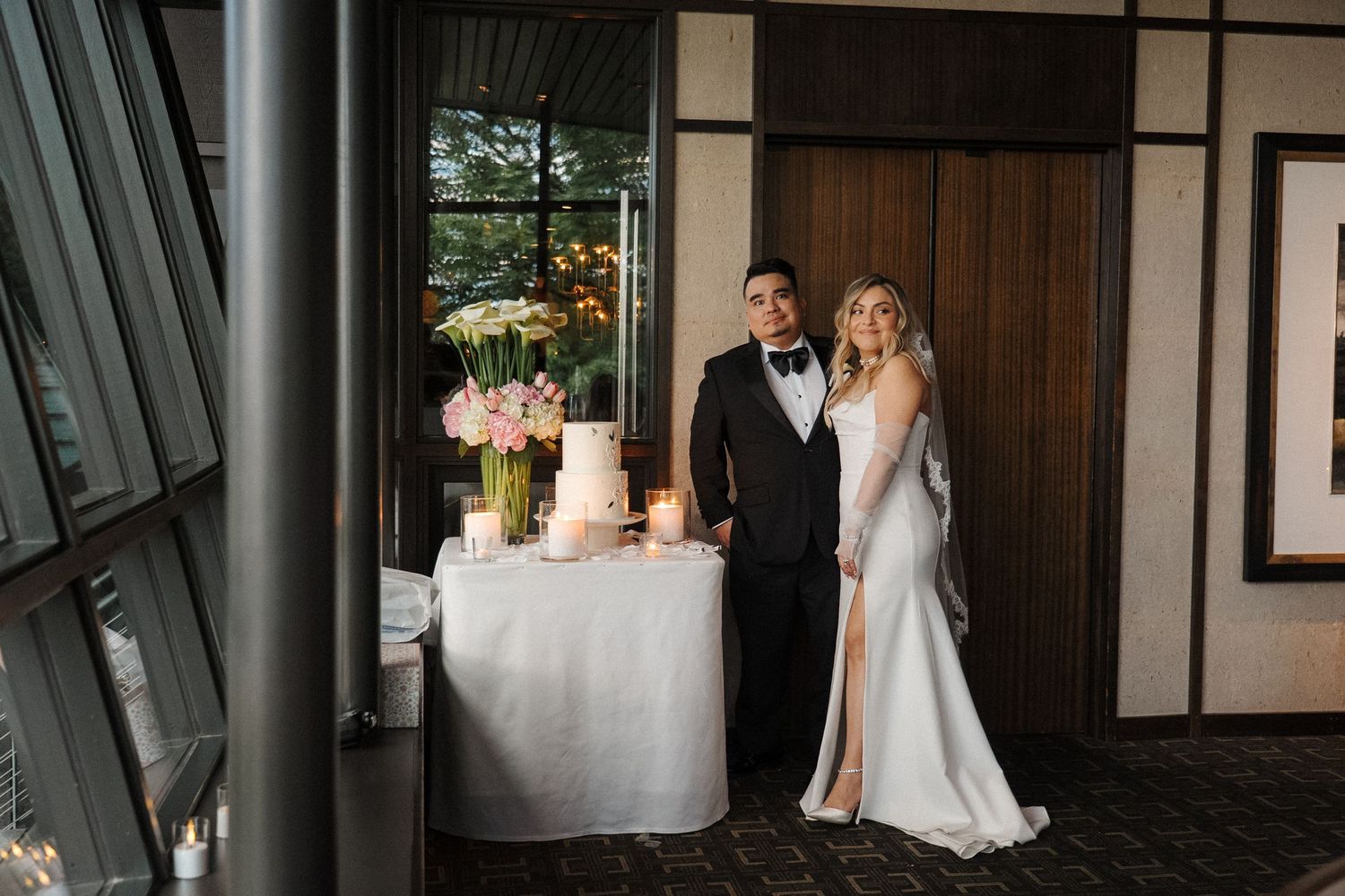 A wedding cake sits elegantly on a wooden table at a rustic venue while dressed in white admire it from afar.