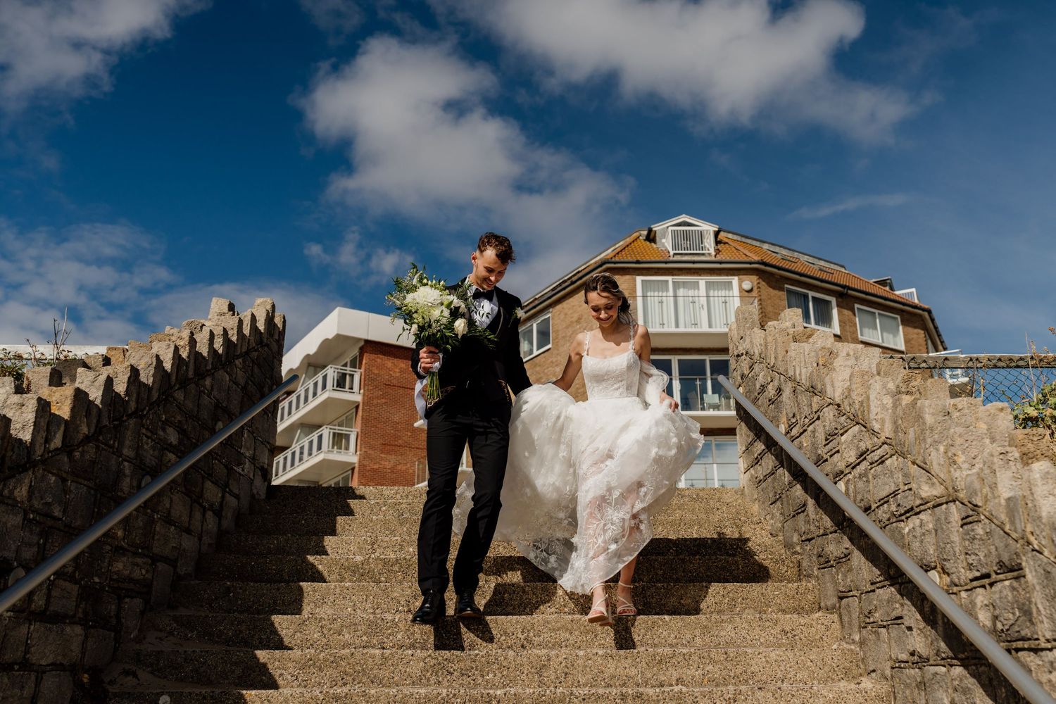 Bride and groom walking down stone steps outdoors on a sunny day, with blue sky and a building in the background.