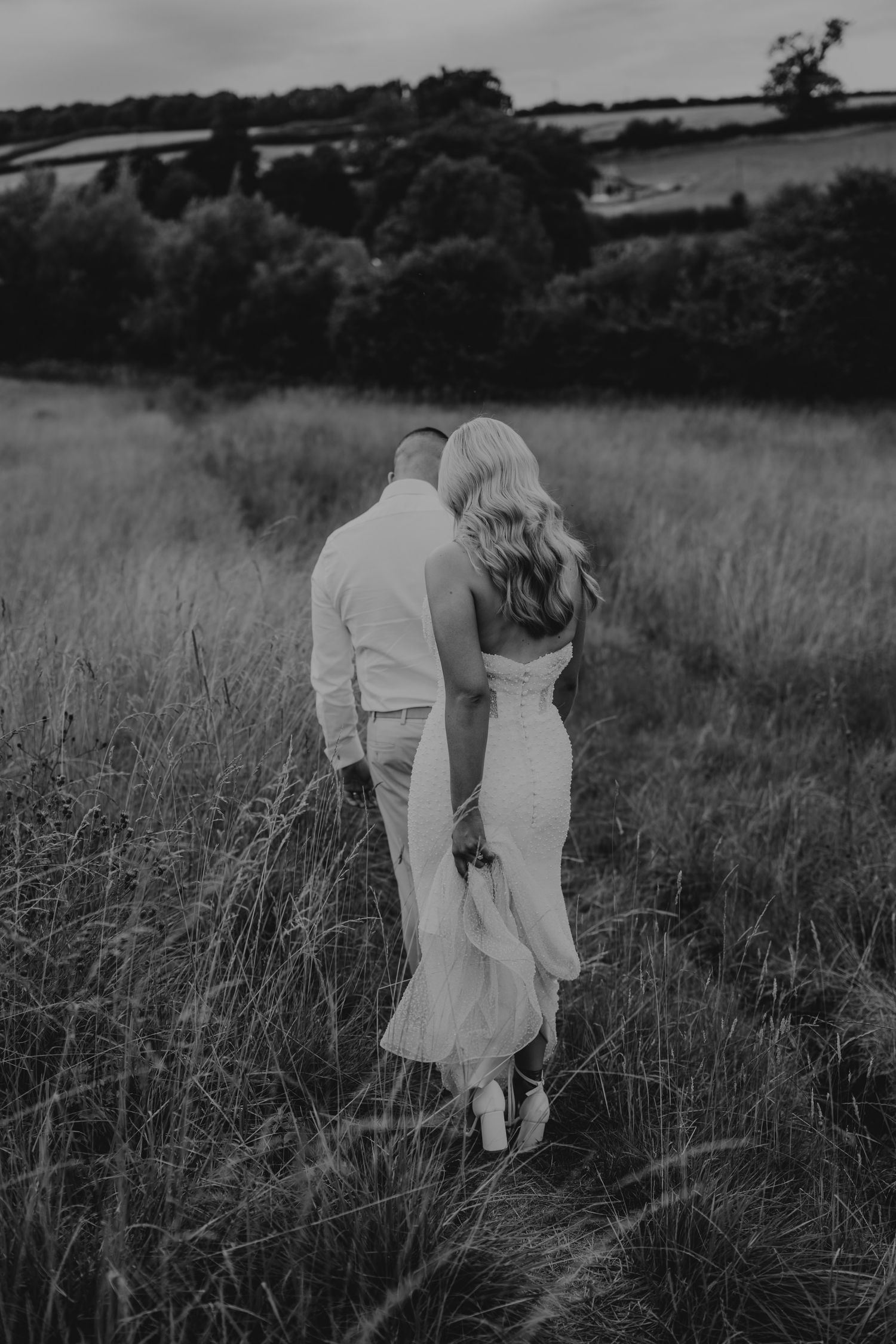 Black and white photo of a couple walking through a meadow, the bride in a flowing dress holding hands with her groom.
