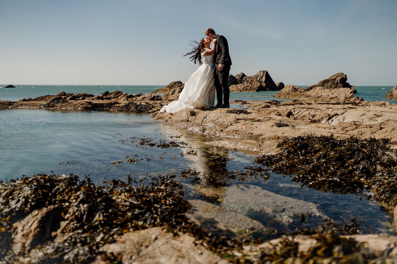 Bride and groom kissing on rocky coastal shoreline with turquoise ocean water and clear blue sky backdrop.