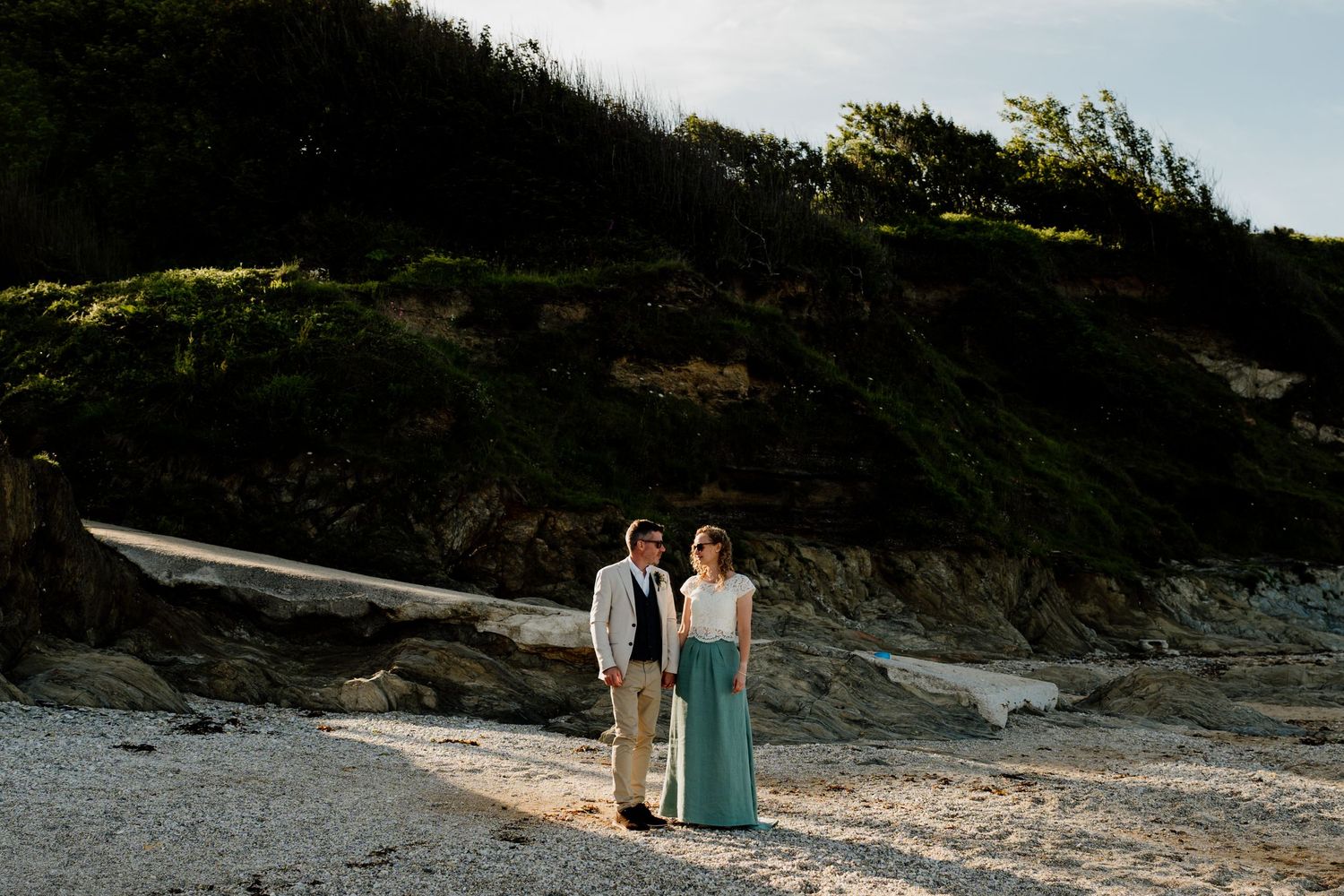 Couple standing on a rocky beach with lush green cliffs behind them during golden hour.