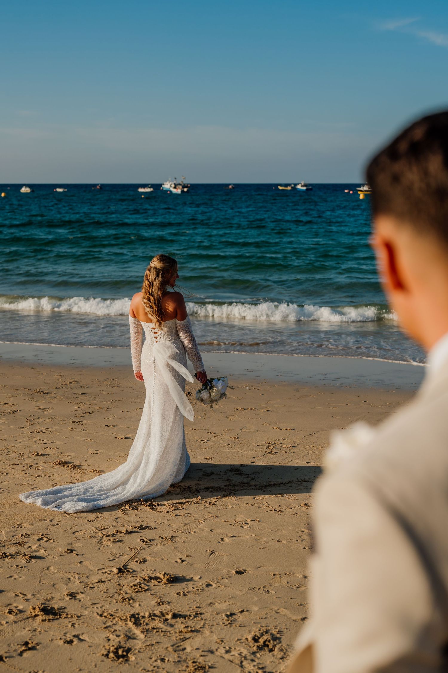 Bride in white off-shoulder gown holding bouquet stands at ocean's edge on sandy beach, groom in foreground.