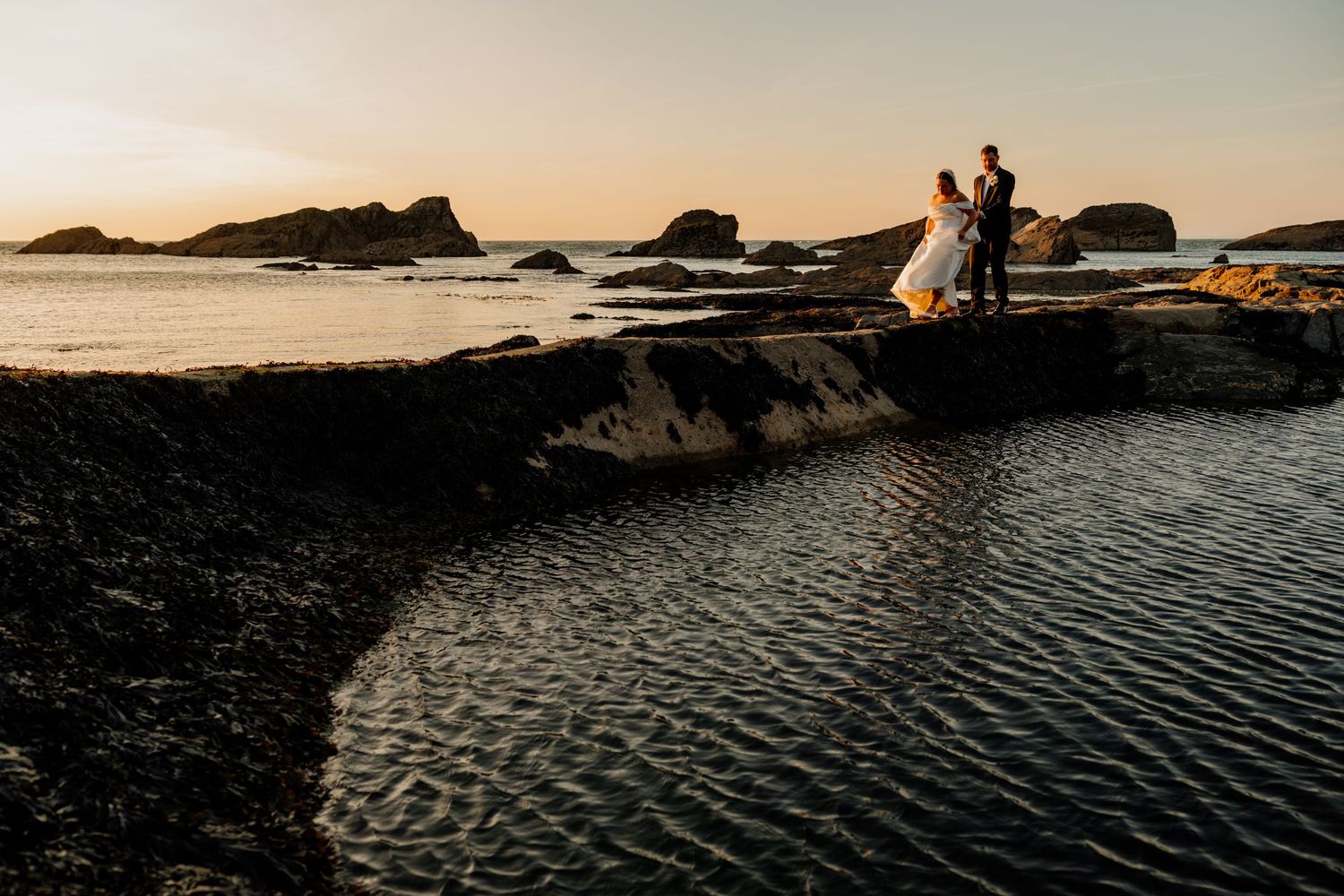 Couple in wedding attire standing on rocky coastal cliffs at golden sunset with ocean waves and rock formations in background.