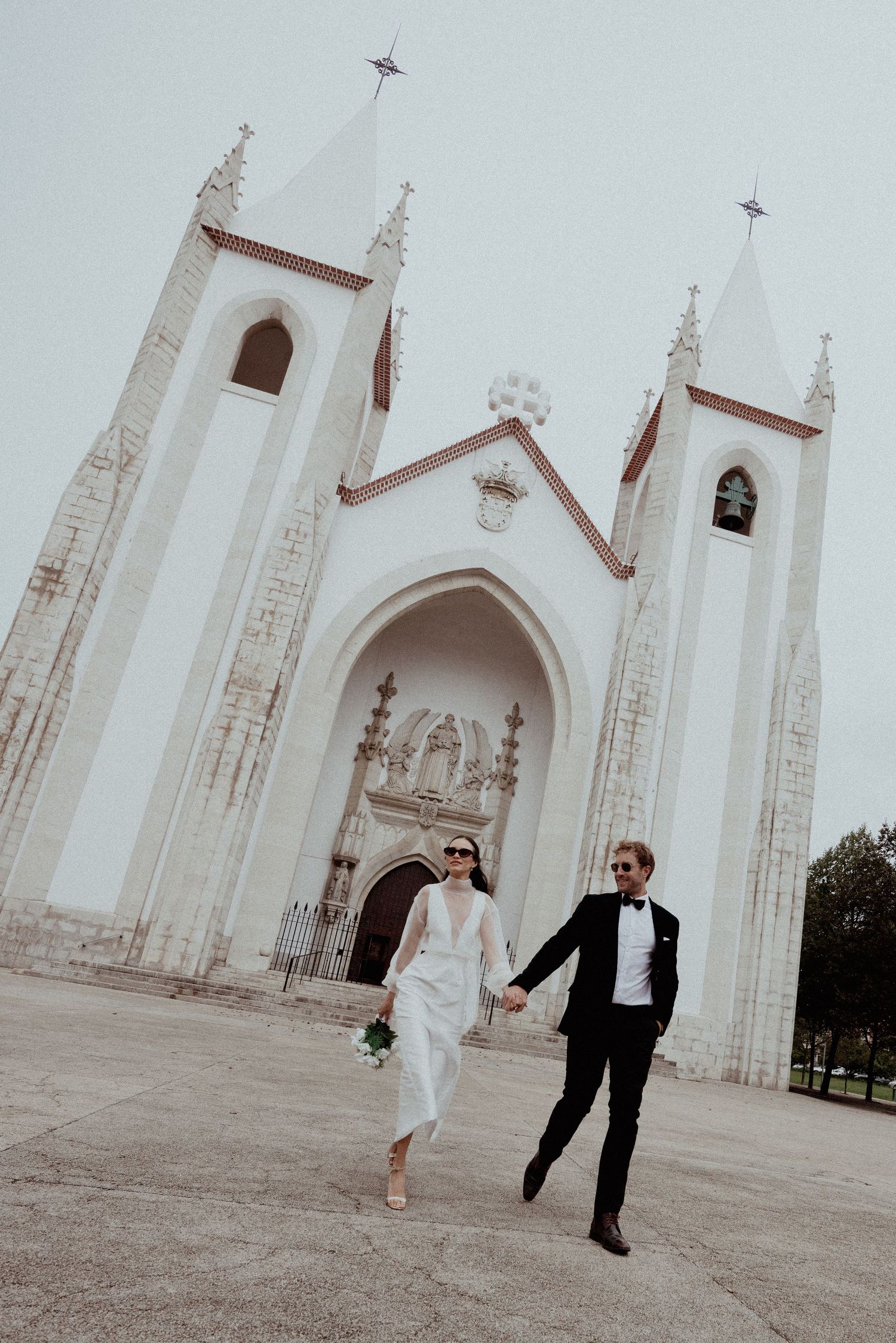 Joyful couple celebrating their wedding day running outside a grand white church with gothic spires.