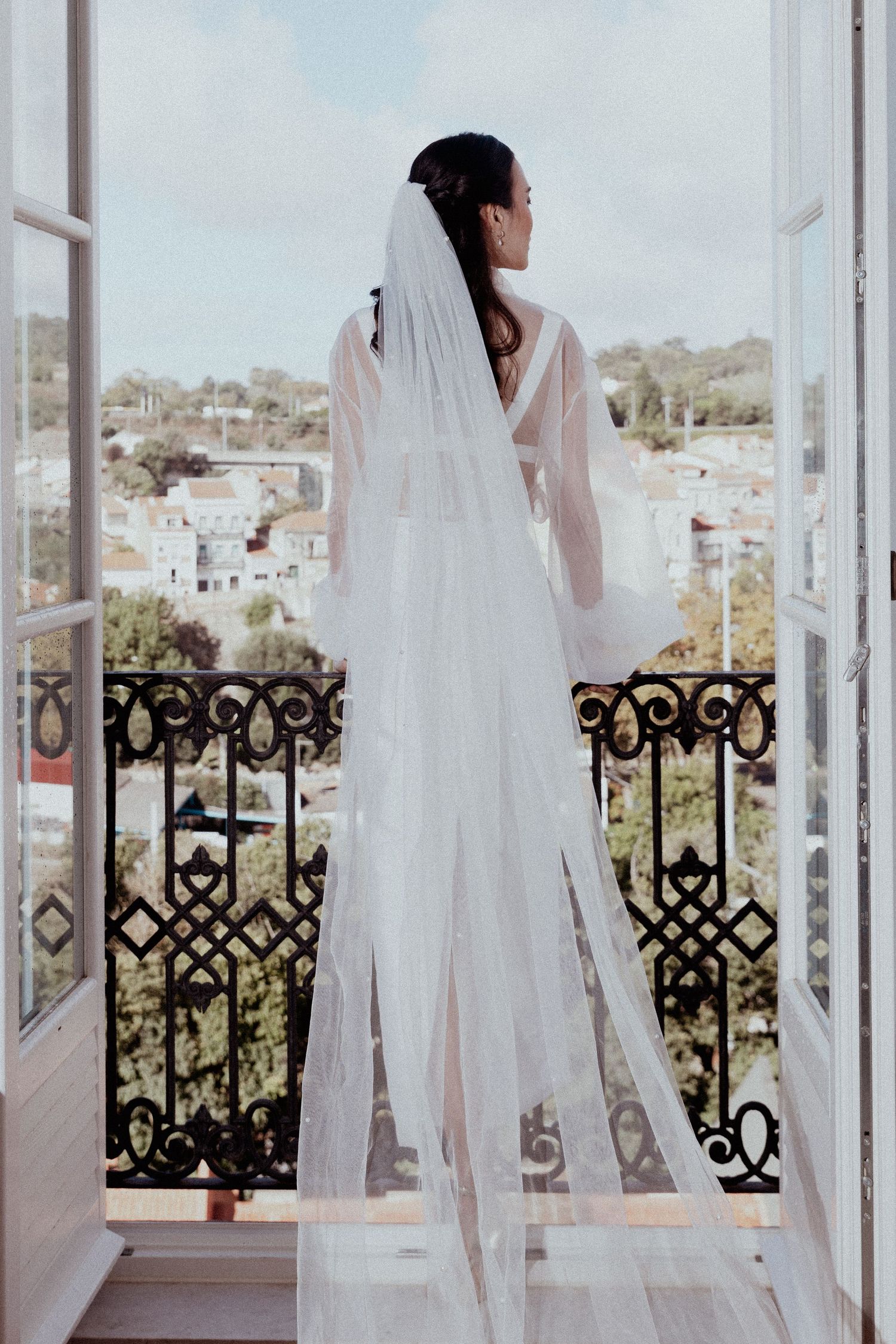 A bride in a flowing white veil and robe stands on a balcony overlooking a scenic Lisbon cityscape through ornate iron railings.