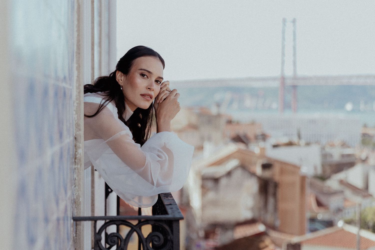 A beautiful bride in white dress looking out over a cityscape with the 25 de Abril Bridge visible in the background of Lisbon, Portugal.