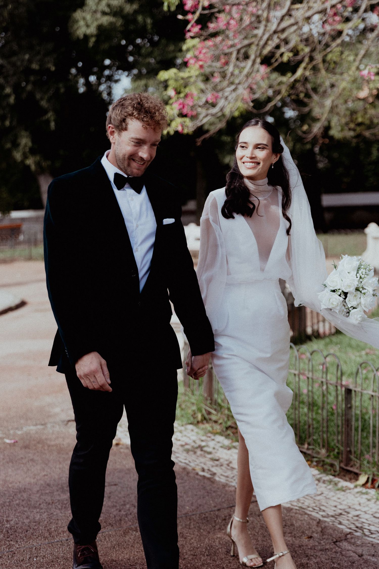 A newlywed couple walks hand in hand outdoors, wearing formal black tuxedo and fitted white wedding dress with veil.