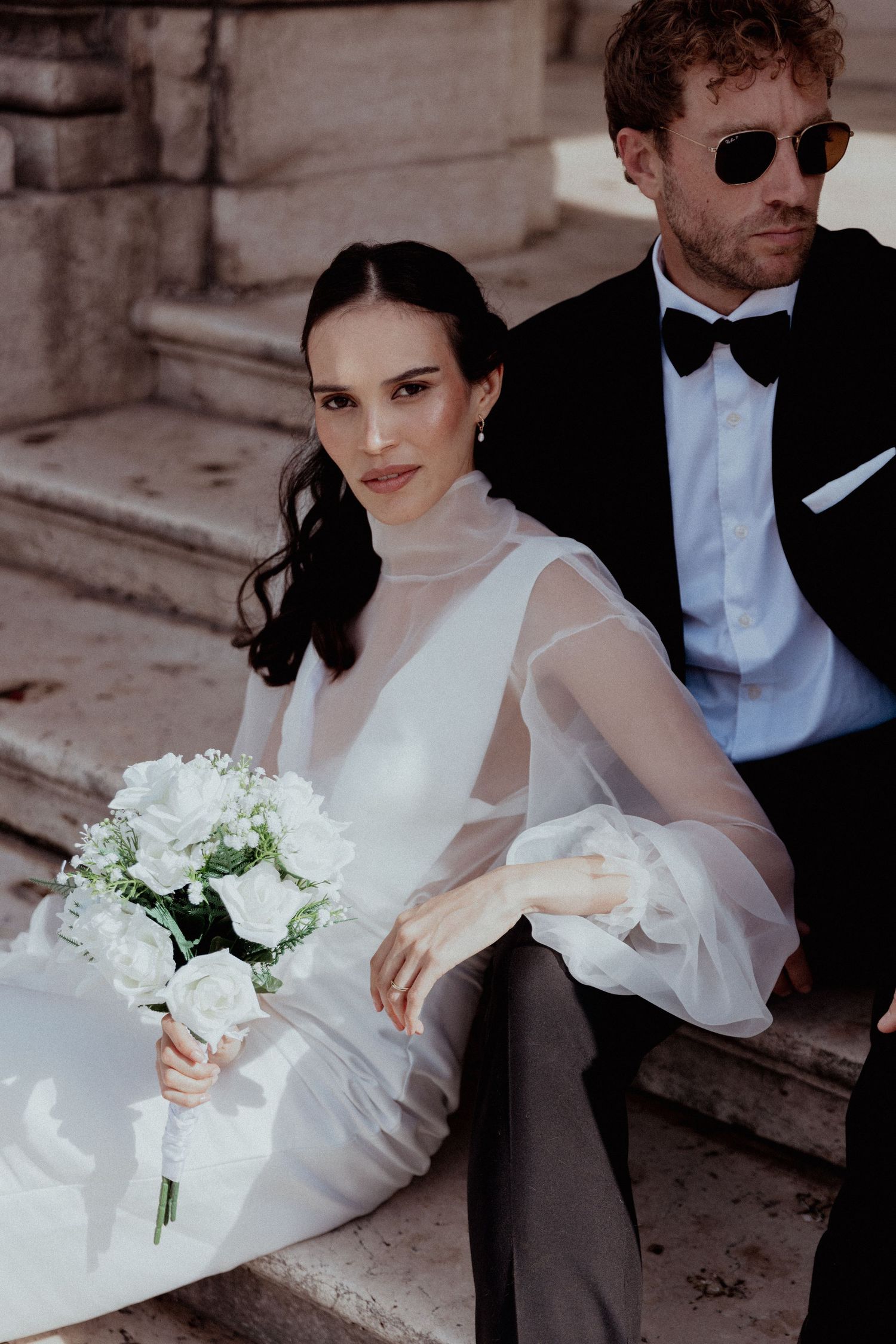 Bride in a flowing white dress and groom in dark suit sit gracefully on stone steps for elegant wedding portraits.