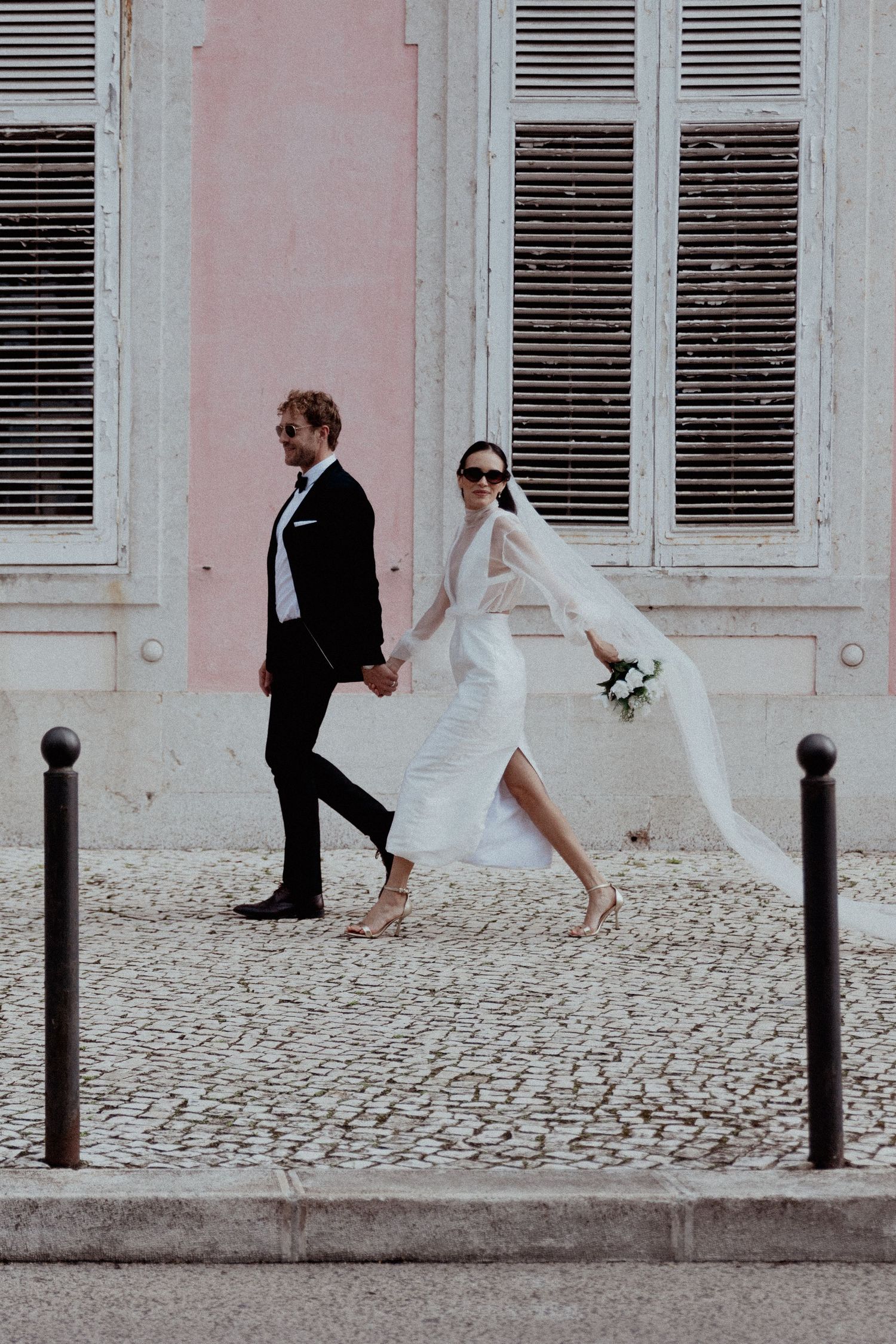 Stylish newlyweds in formal attire walking hand in hand along a cobblestone street beside a pink wall with white shutters.