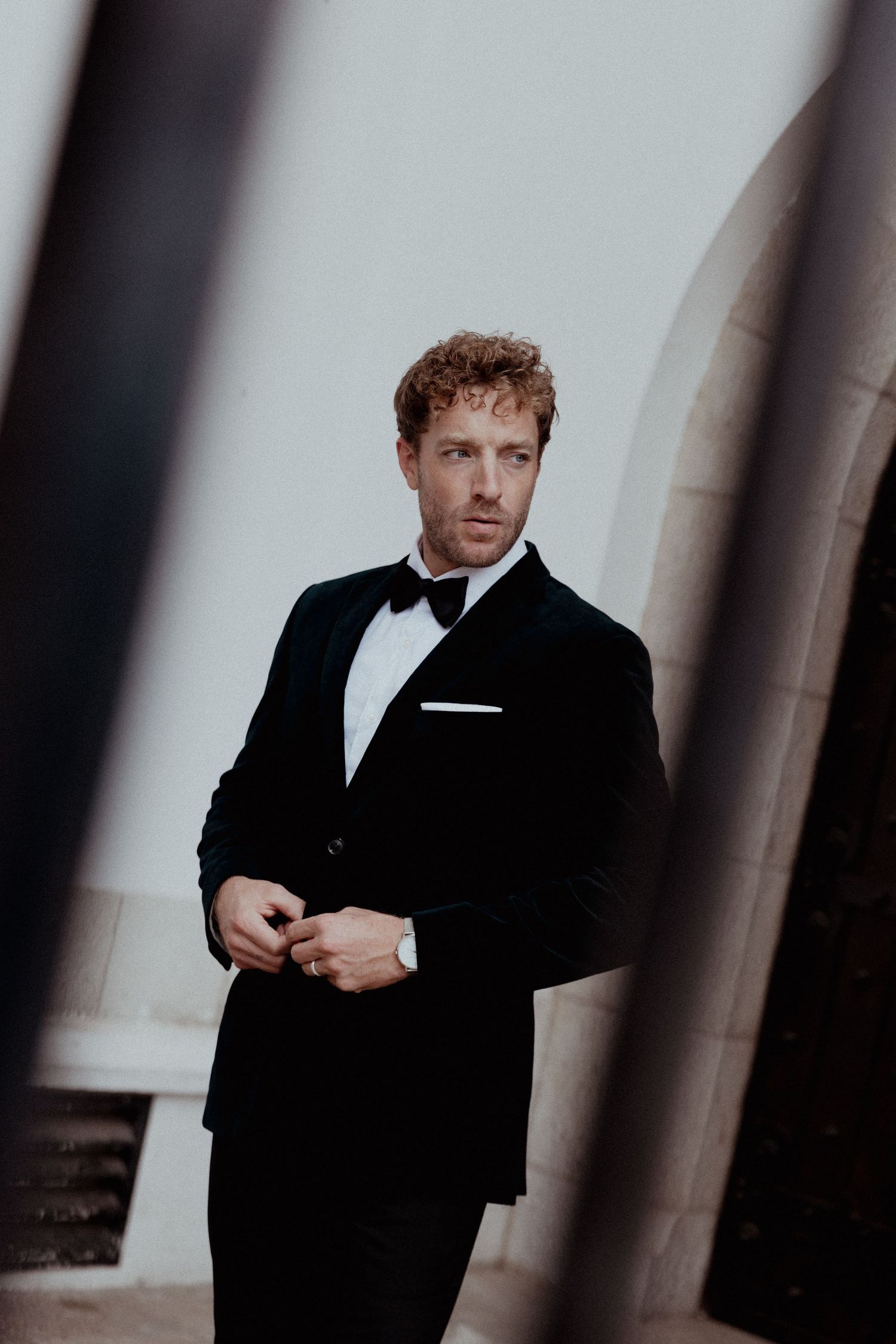 Elegant groom in classic black tuxedo and bow tie poses against column in moody architectural setting.