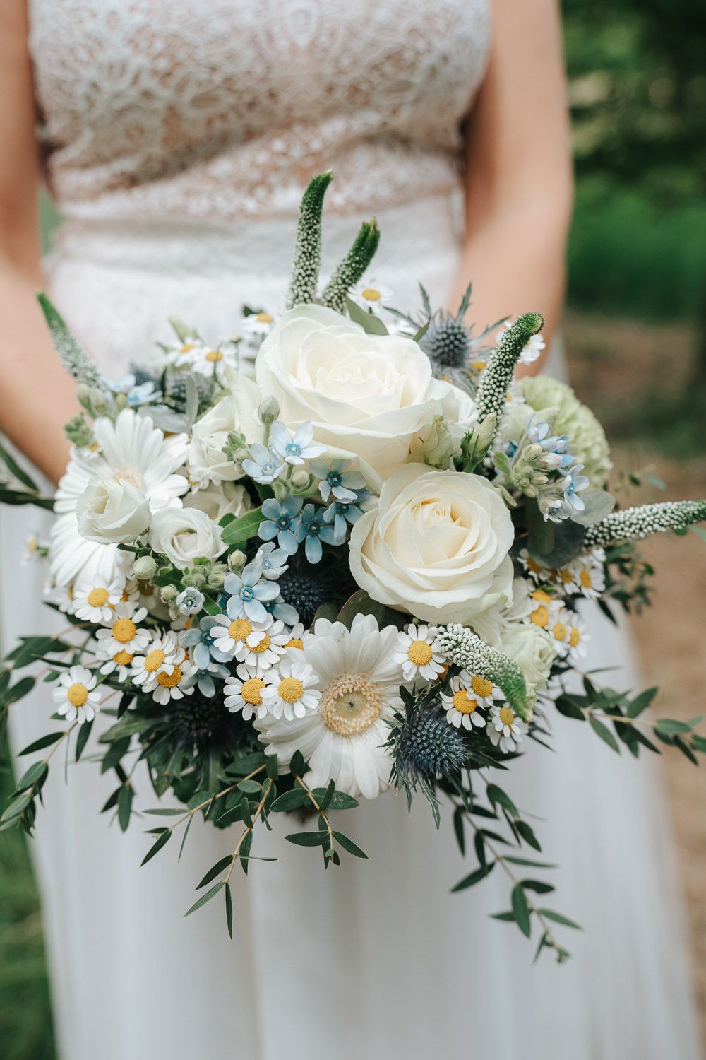 A romantic wedding bouquet featuring white roses, daisies, and blue thistle flowers against a white dress background.