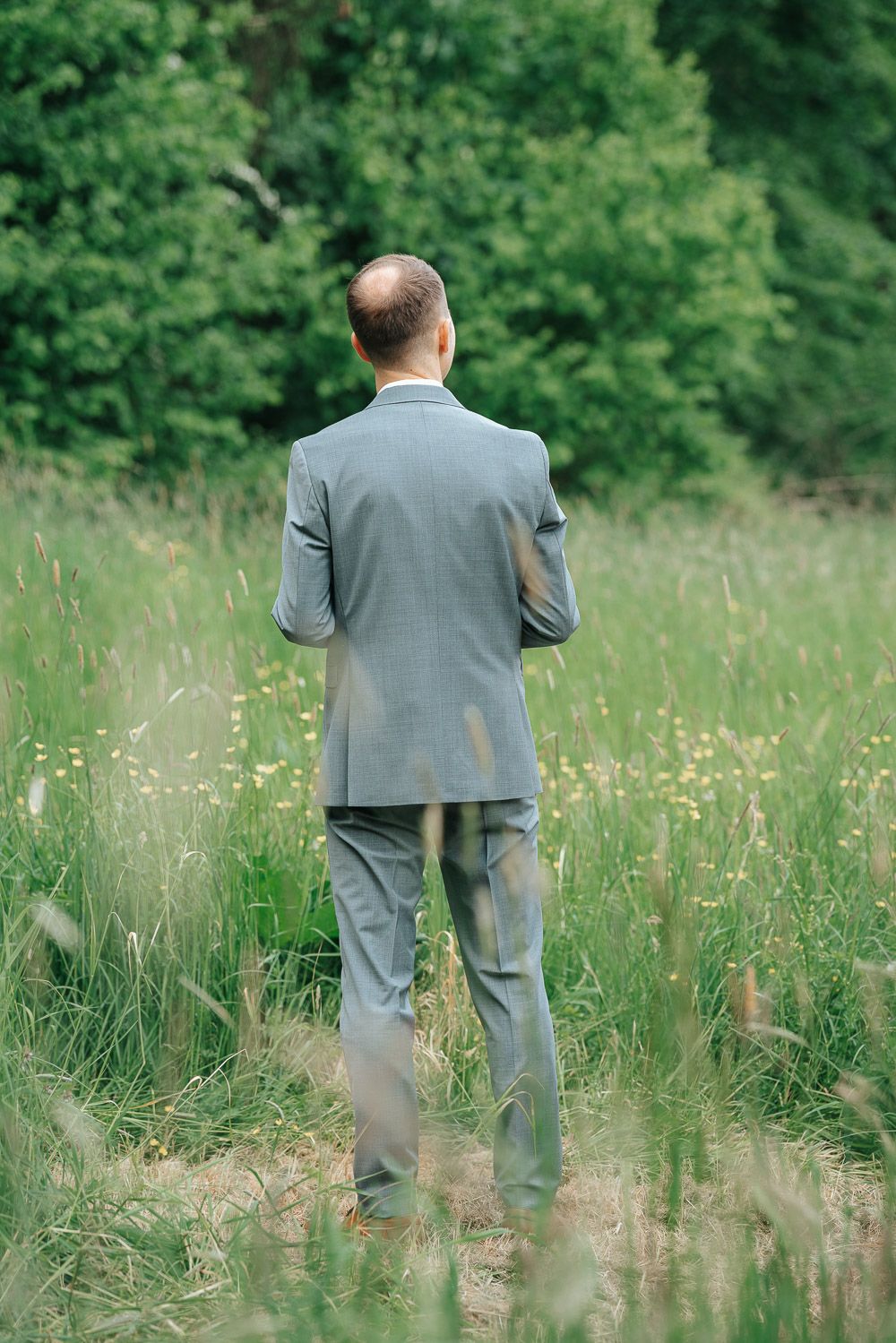 A person in a gray suit stands alone in a summer meadow with tall grass and wildflowers on a sunny day.