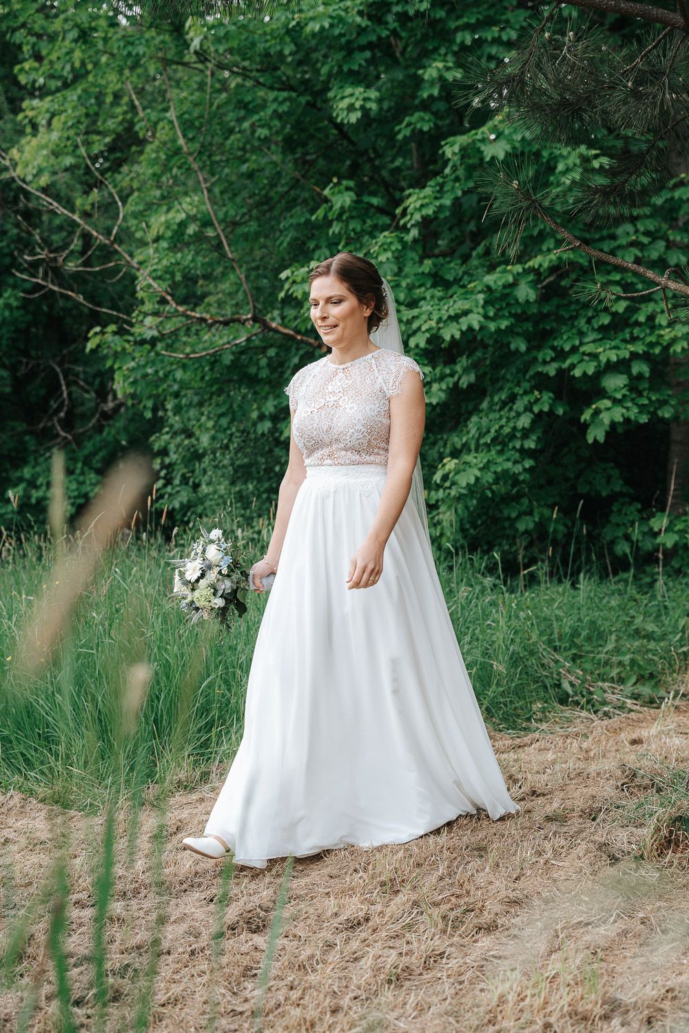 A bride in a lace-topped white wedding gown holds a bouquet while standing in a scenic wooded outdoor setting.