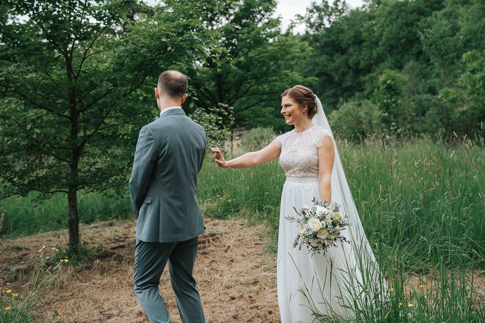 A couple shares their first look moment on a woodland trail, with the bride in white holding a natural bouquet.