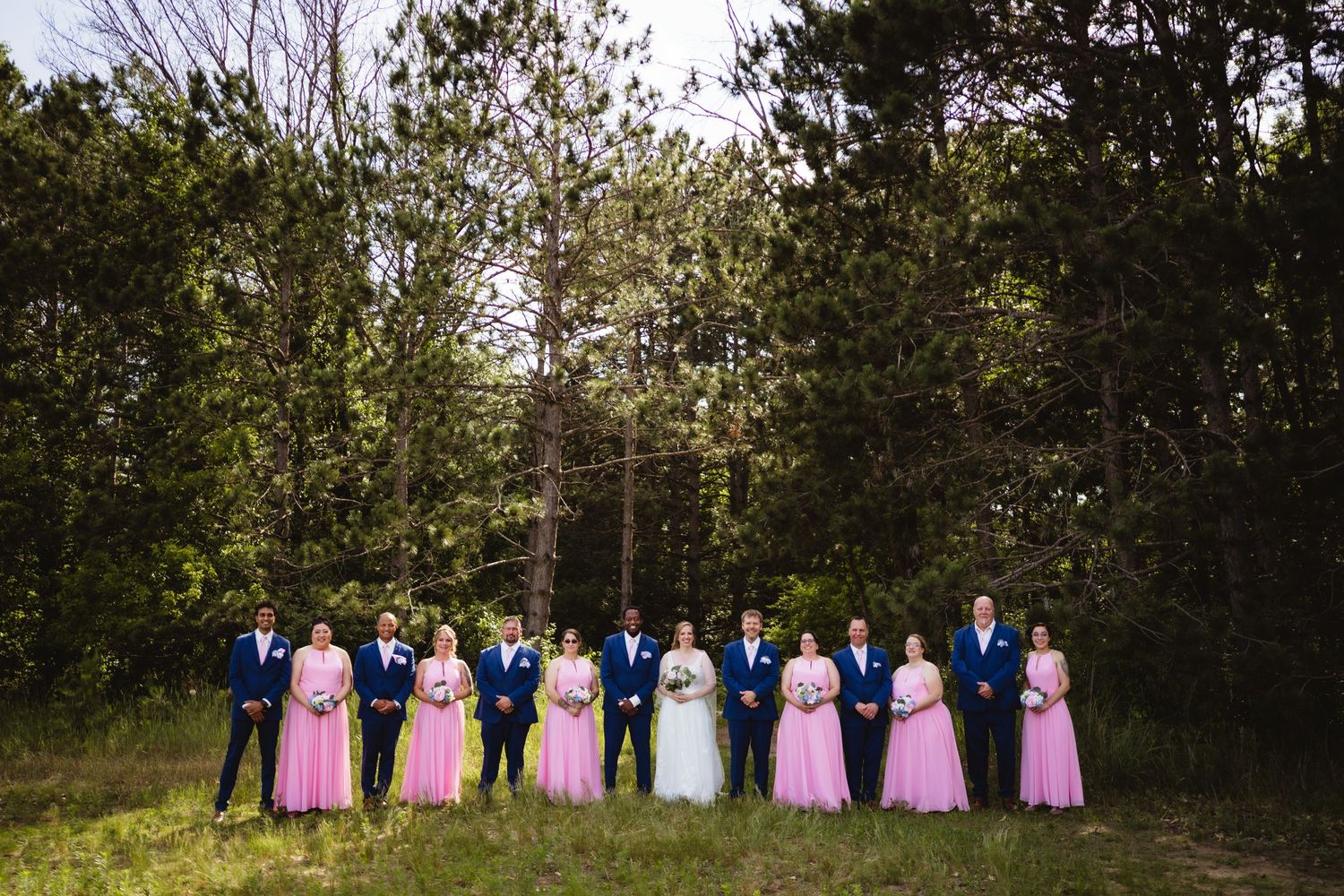 A group of wedding party members in pink and blue dresses stand in a row against a wooded outdoor backdrop.