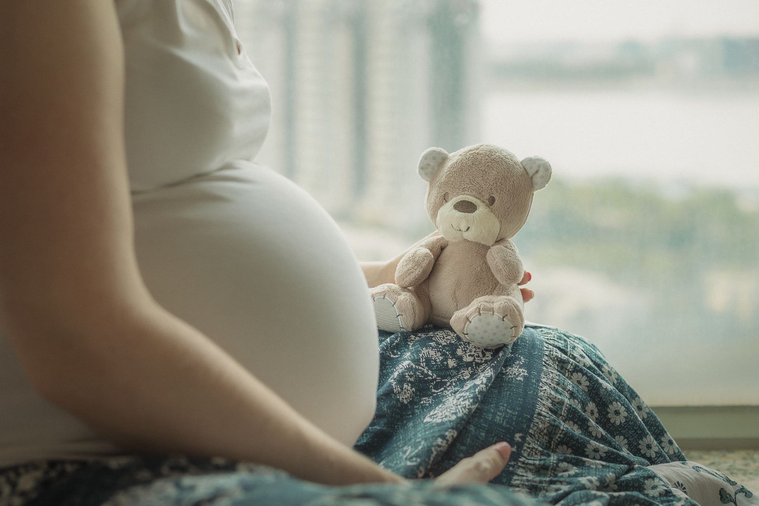 Pregnant woman in white tank top folding baby clothes while sitting by window with natural light.