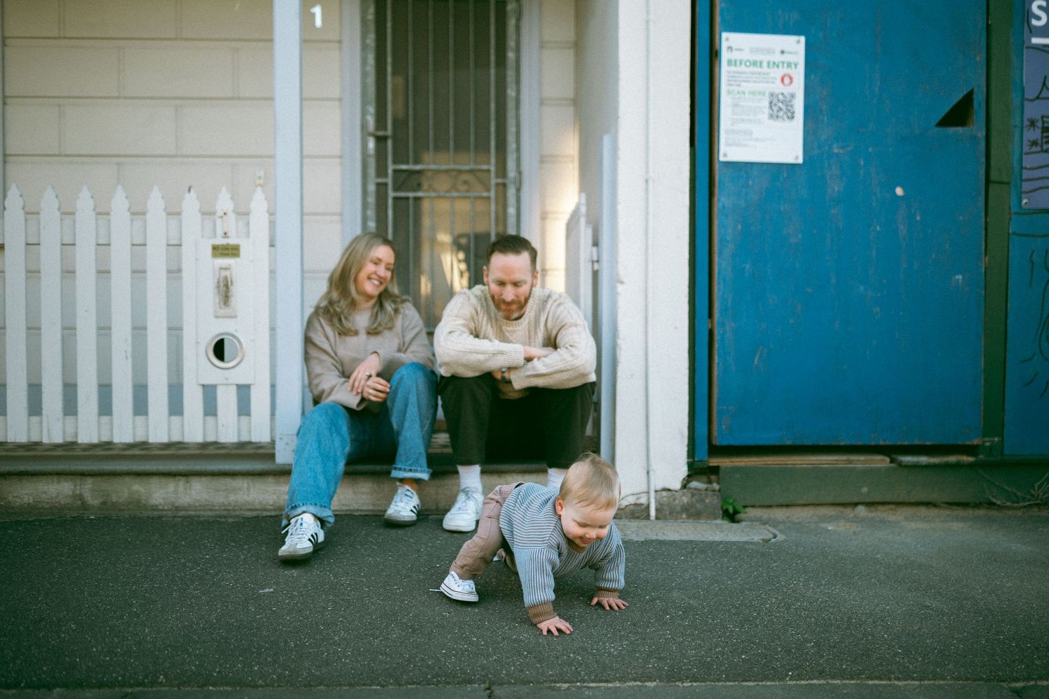 A family sit relaxed on their front door step in Richmond while their small toddler plays on the ground in front of them.