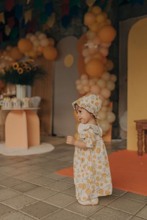 Small child wearing floral print dress and bonnet standing at a decorated party venue with balloon garlands.