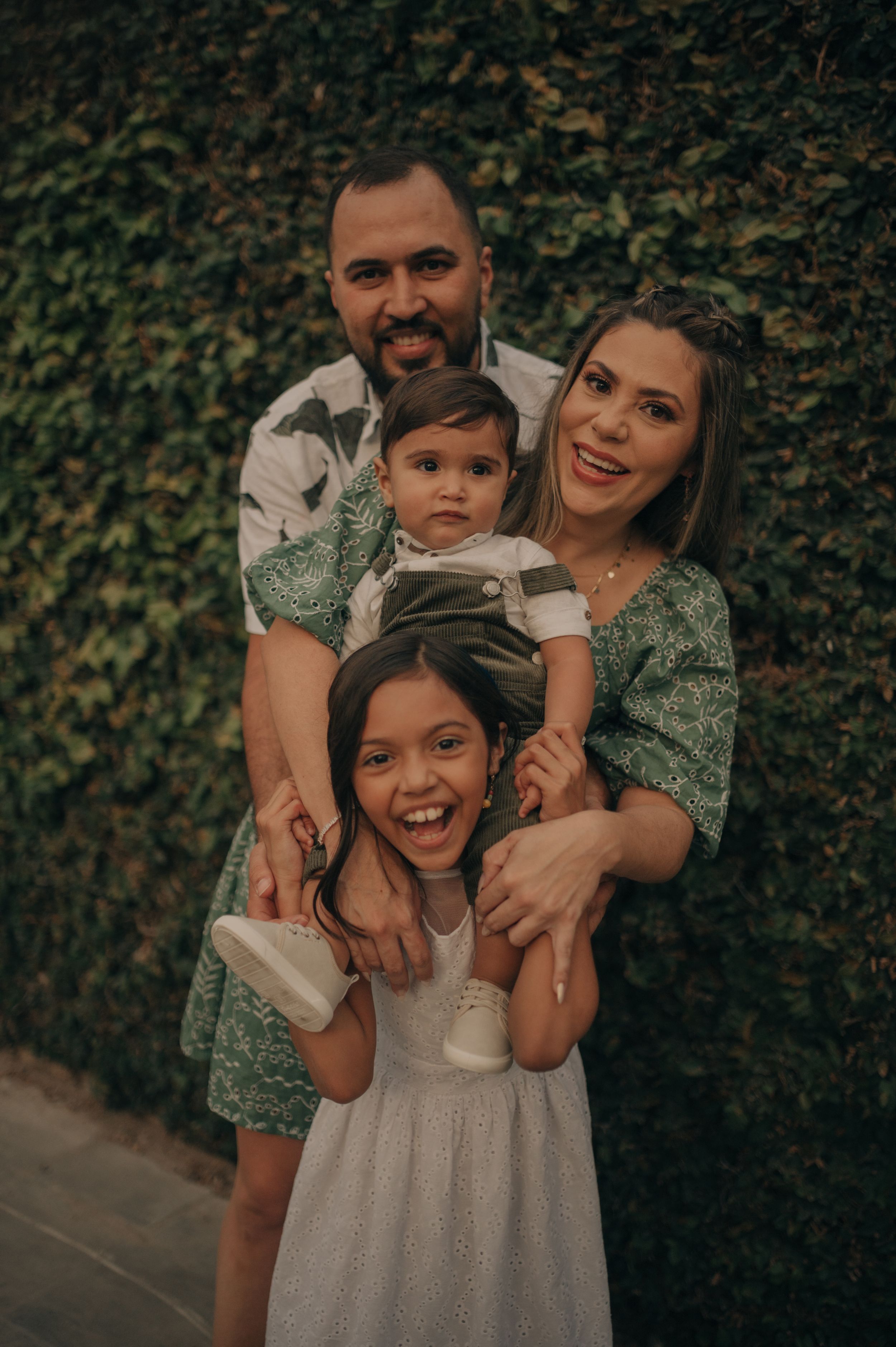 A family wearing coordinated green outfits poses together against an ivy-covered wall.