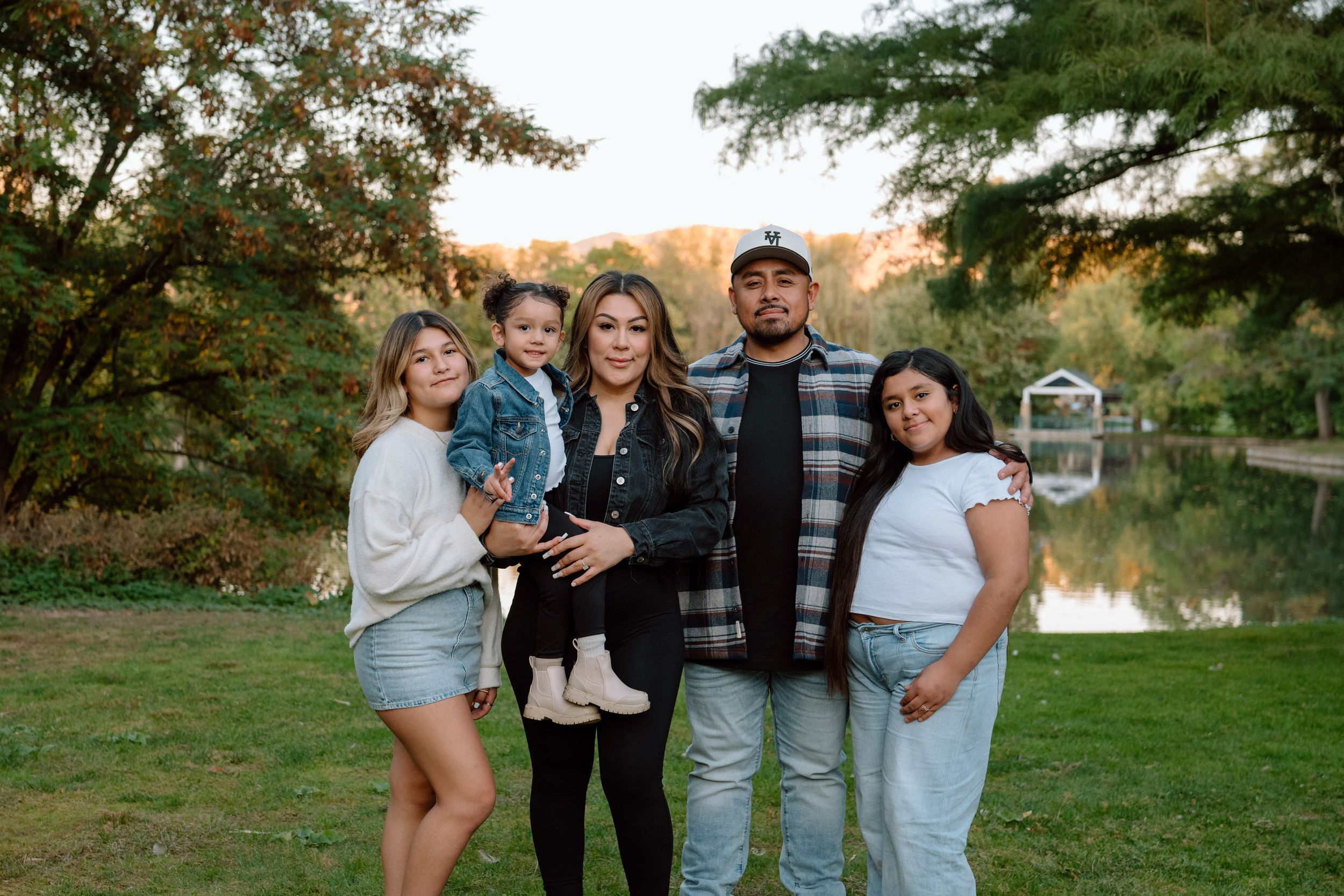 A family stands together outdoors in casual attire by a lake during golden hour with trees in the background.