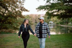 A couple holding hands while walking along a serene pond surrounded by autumn trees in a peaceful park setting.