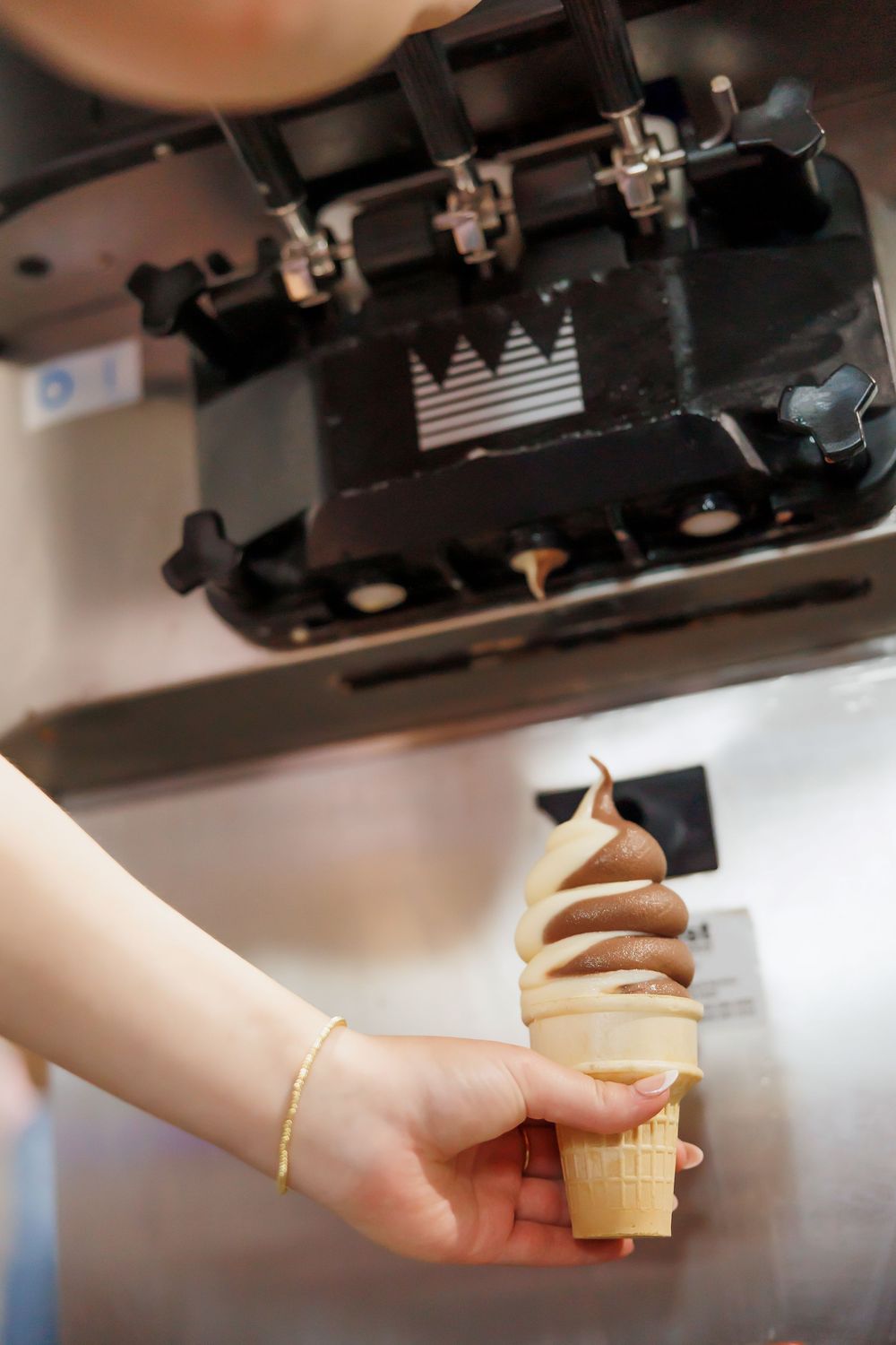 Close-up view of hands dispensing soft serve ice cream from a commercial machine into a waffle cone.