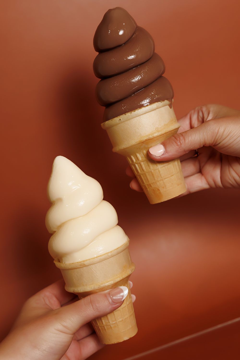 Two hands holding soft serve ice cream cones with chocolate and vanilla swirls against a brown background.