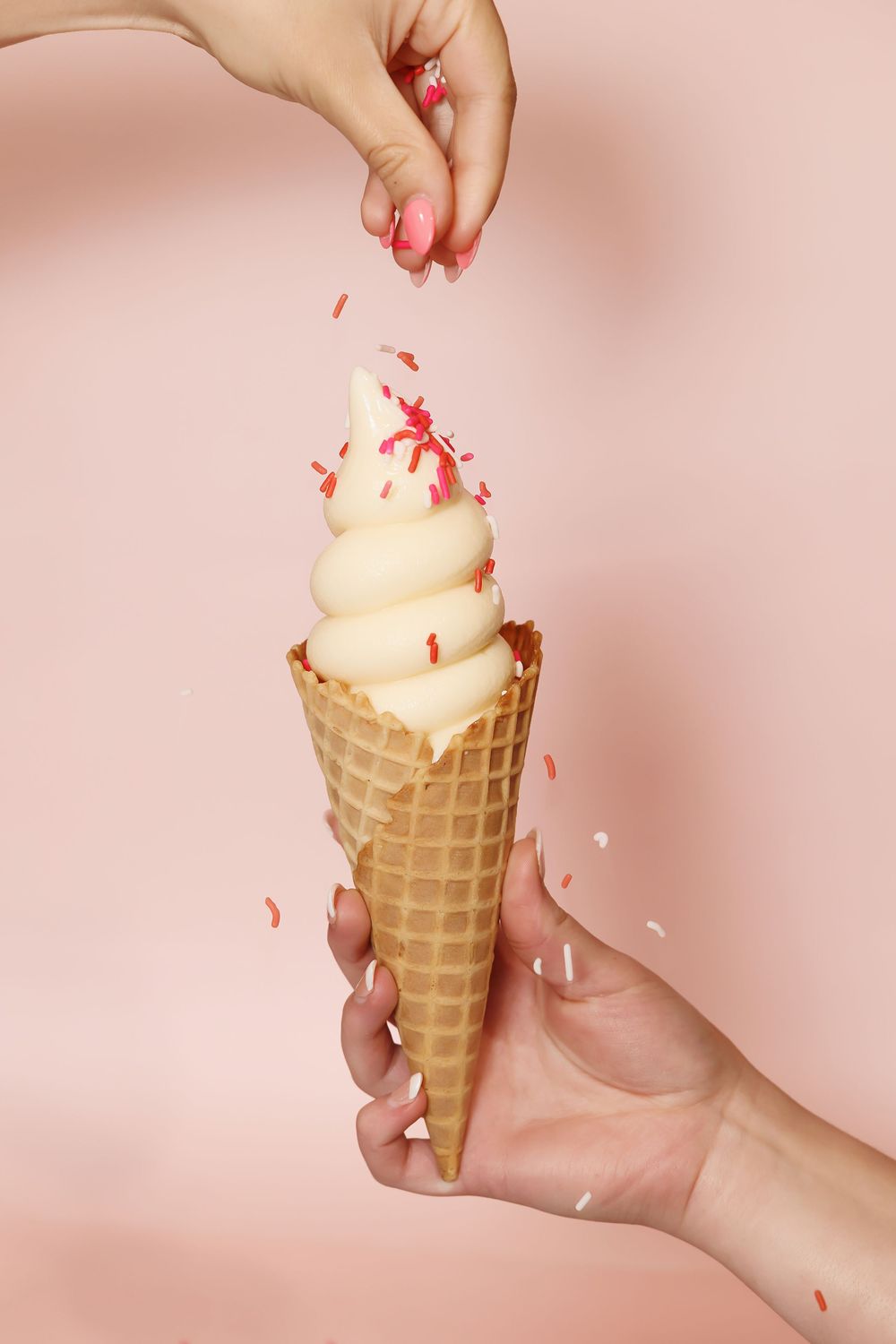 Vanilla ice cream cone with red sprinkles being passed between hands on pink background.